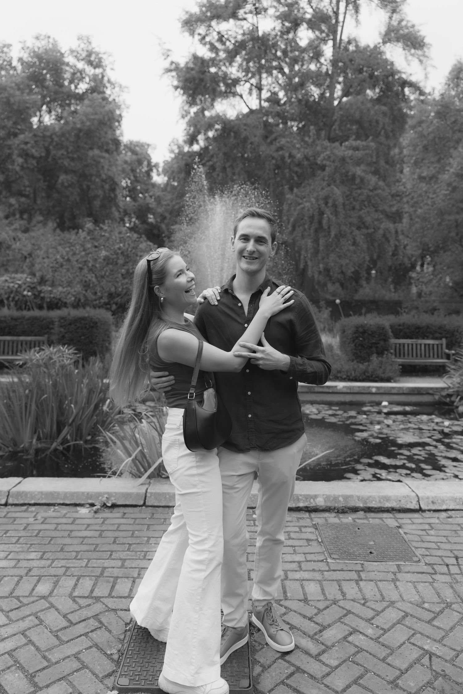 Black and white photo of a happy couple embracing in front of a garden fountain surrounded by lush greenery.