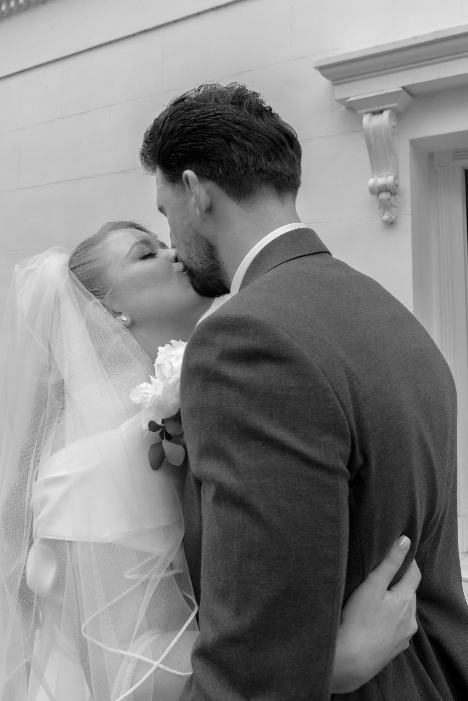 Black and white photo of bride and groom sharing romantic kiss outside, bride wearing veil.