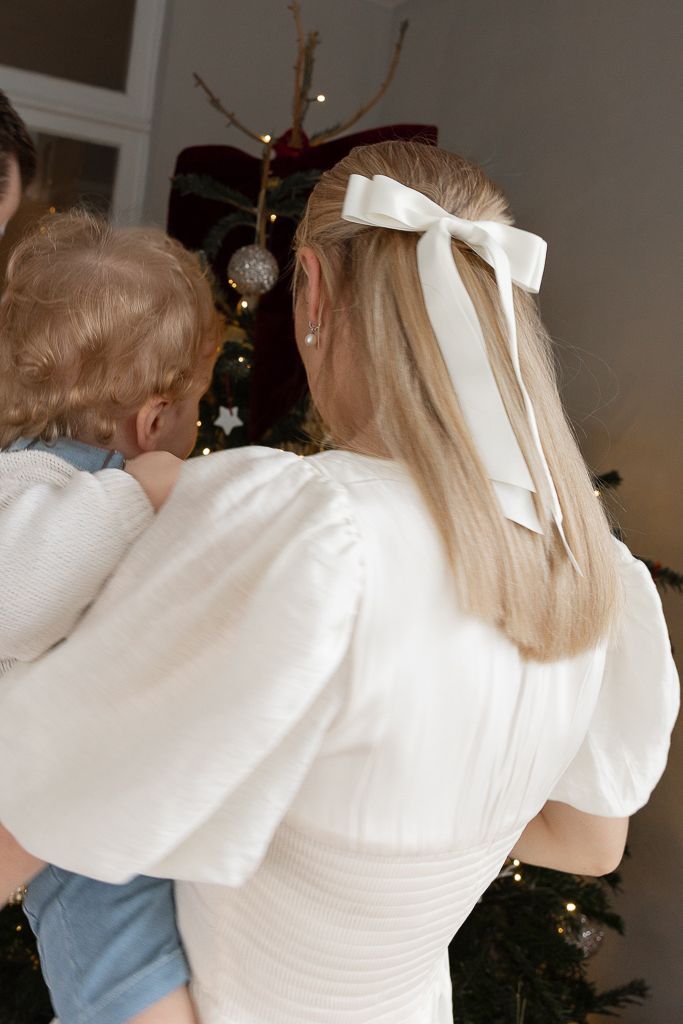 A white bow adorns blonde hair as someone holds a child near a Christmas tree.