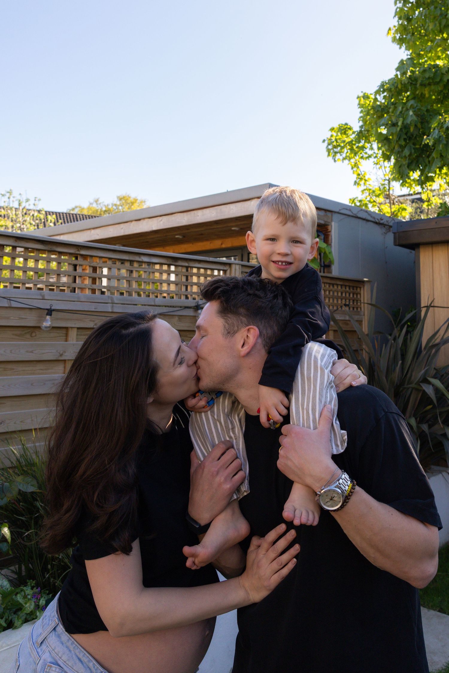 Couple kissing while man holds smiling toddler on shoulders in a sunny outdoor garden setting.