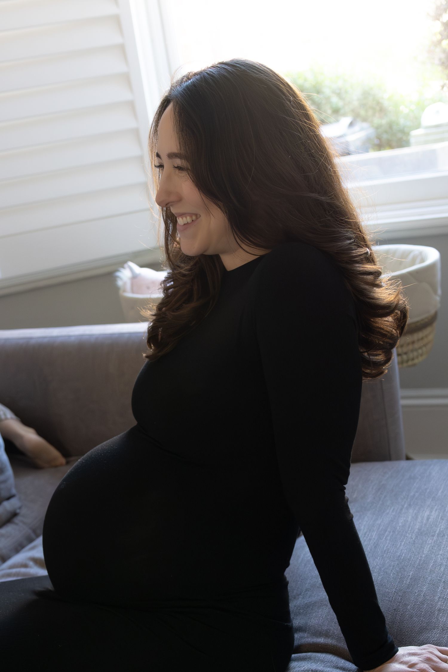 Smiling pregnant woman in black long-sleeve dress sitting on couch in bright home interior.