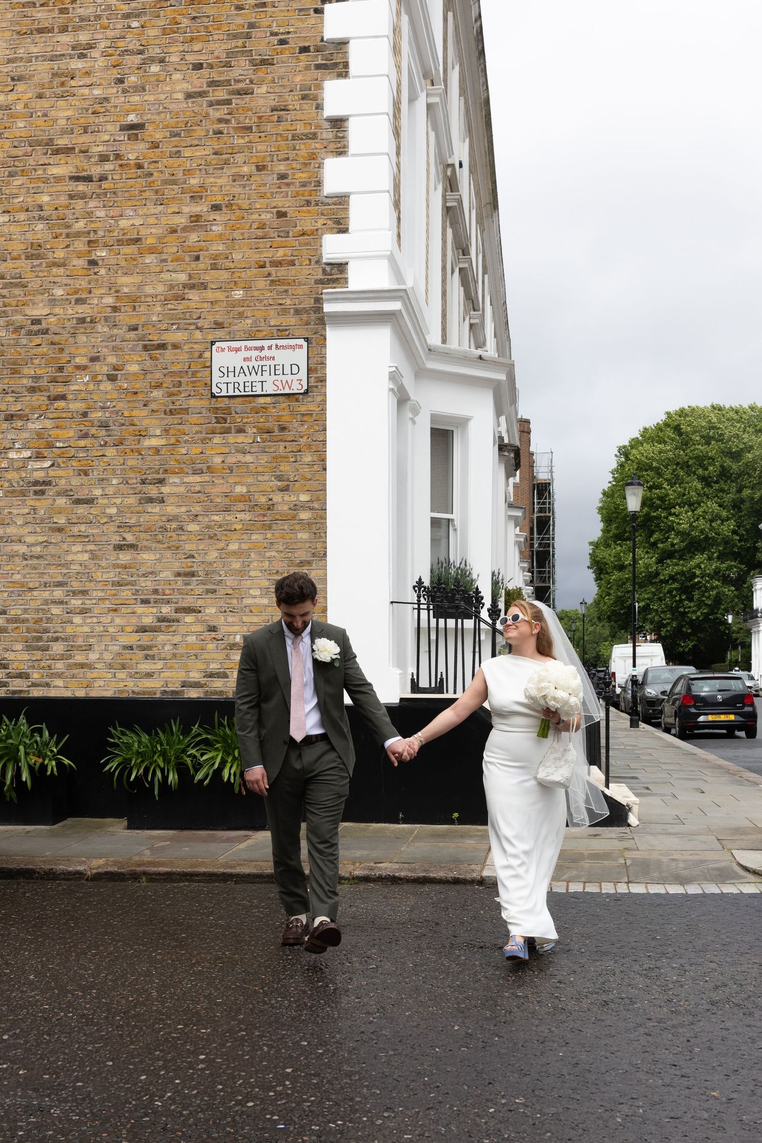 Bride and groom hold hands walking on a wet London street near Shawfield Street sign on a cloudy day.