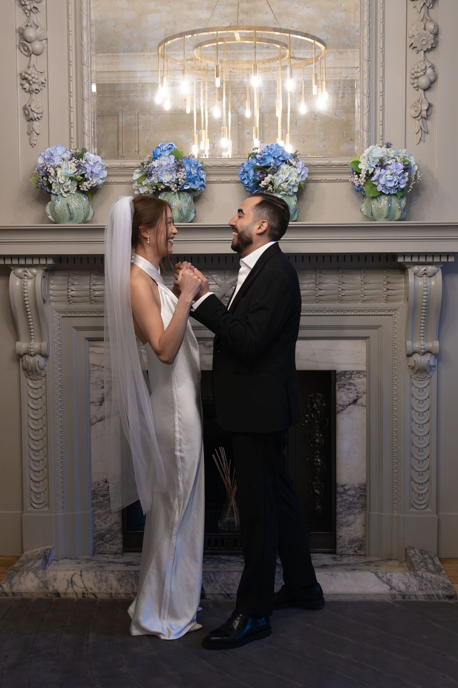 Bride and groom holding hands smiling before ornate fireplace with blue hydrangeas and gold chandelier.