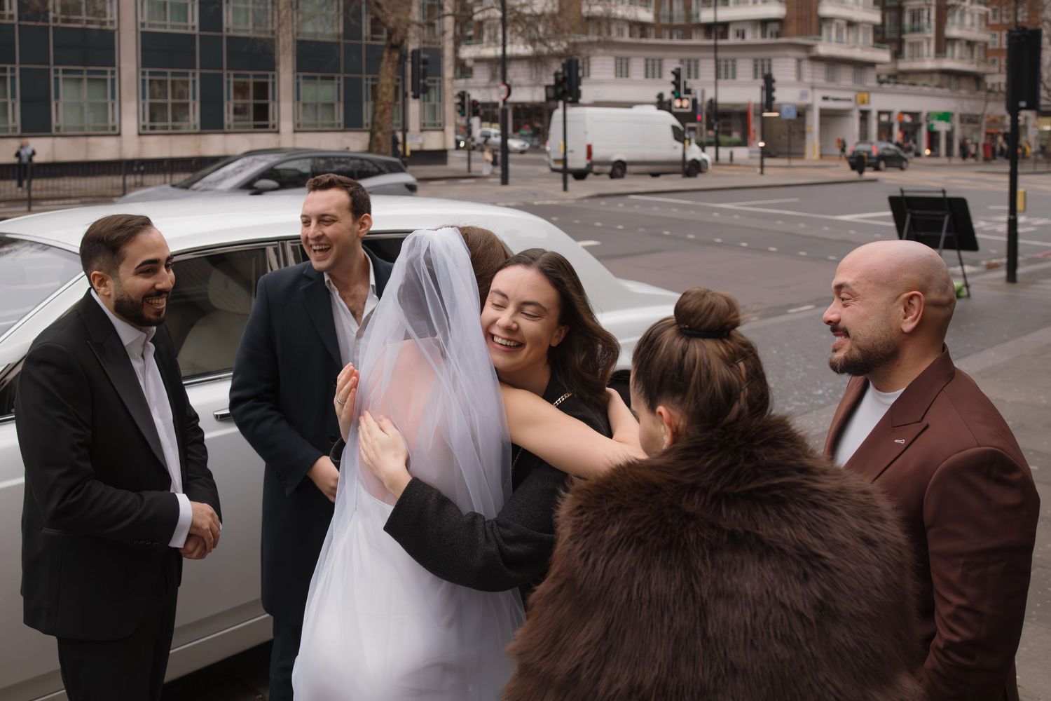 Bride in white dress and veil hugging guests beside a limousine on a city street on her wedding day.