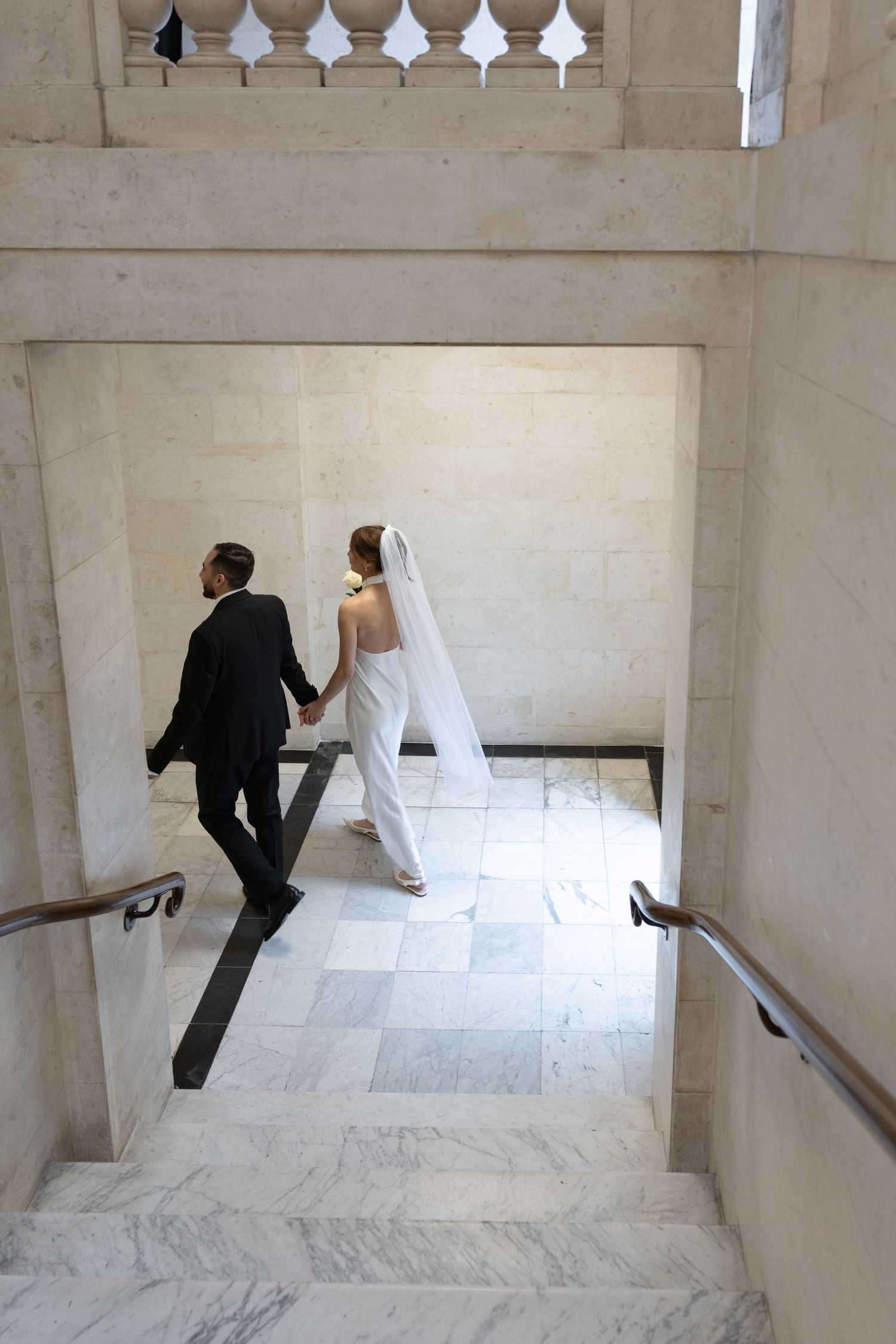 Bride and groom holding hands while walking down marble stairs in wedding attire.