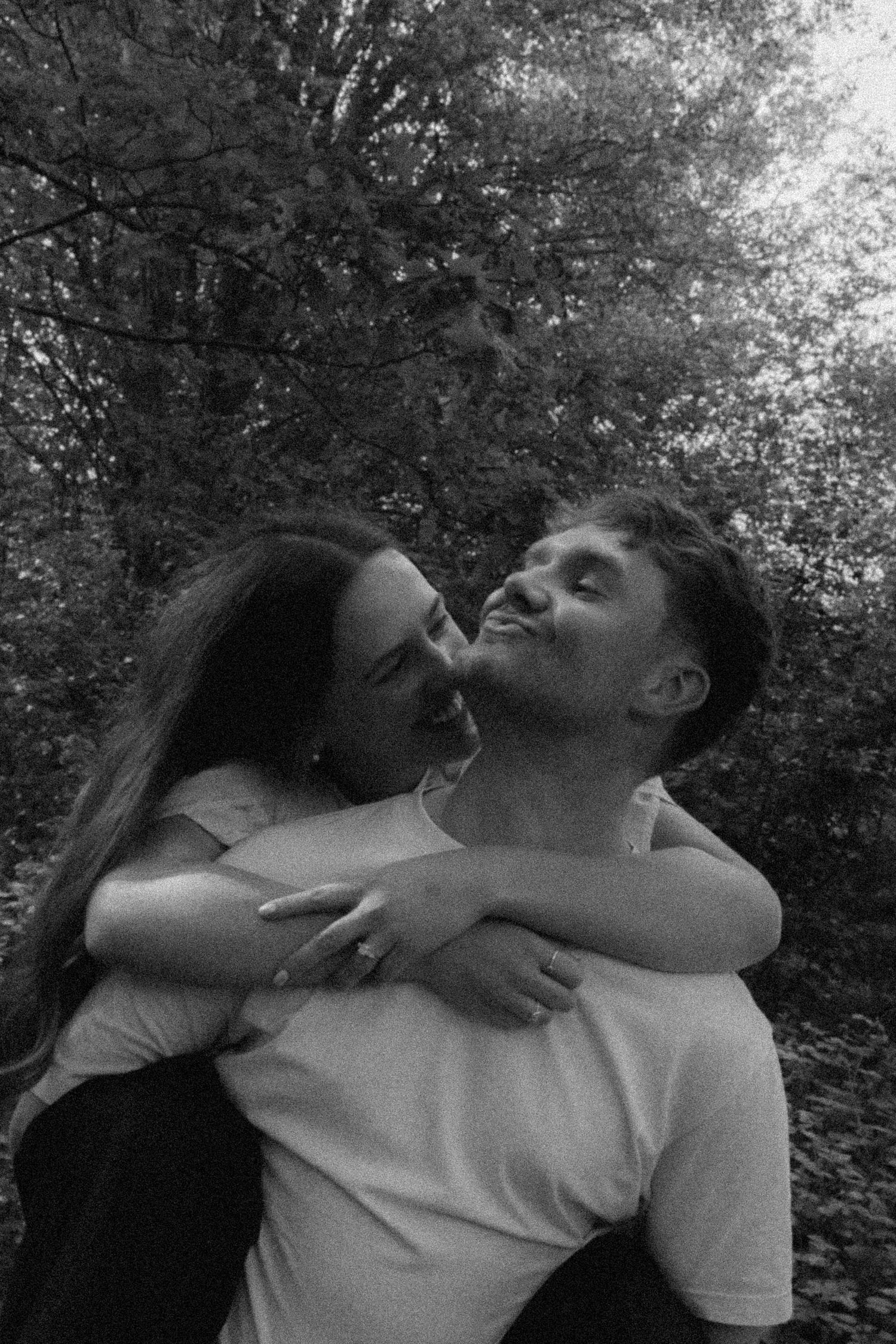 Black and white photo of a couple embracing and sharing a tender moment outdoors among lush trees.