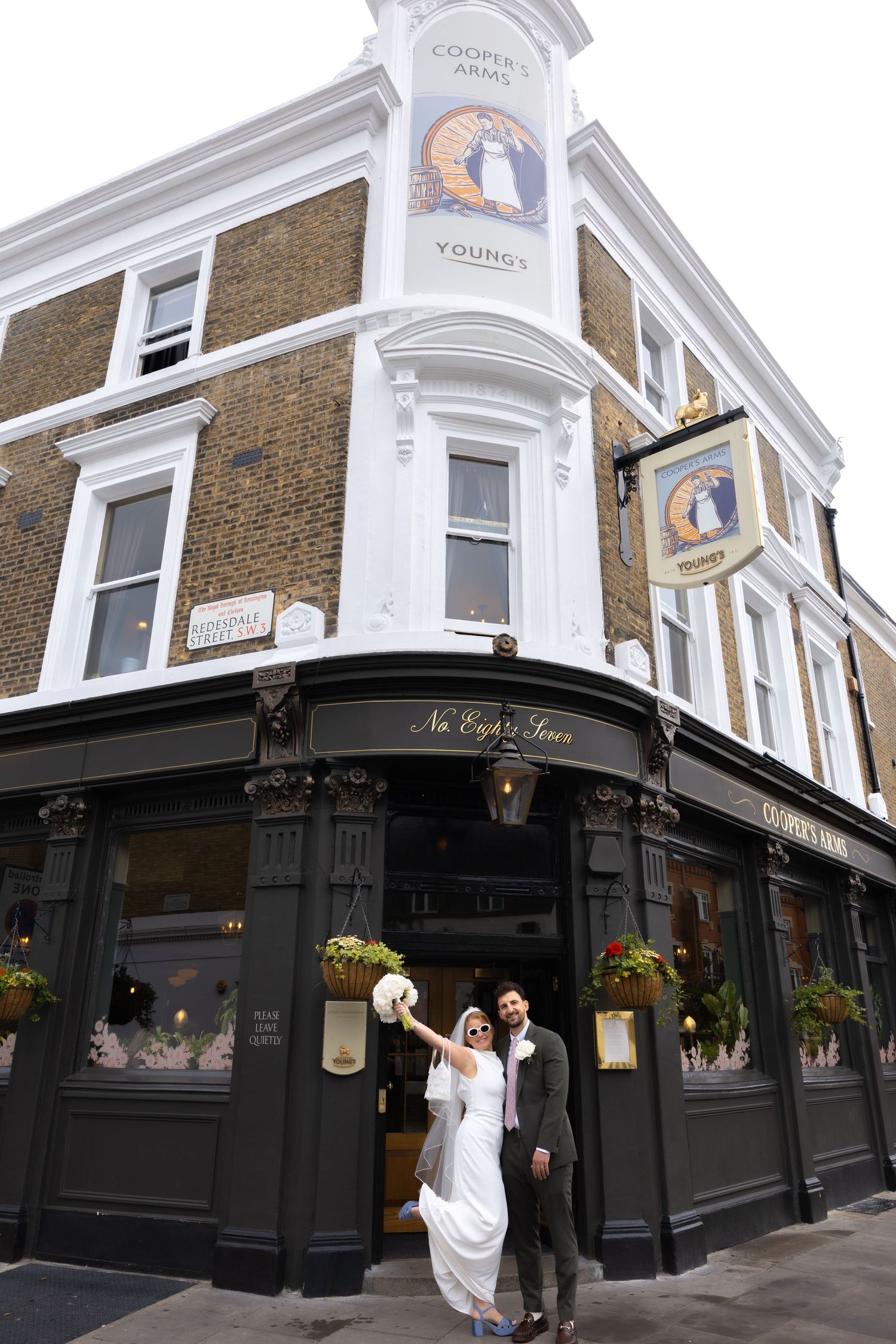 Bride and groom celebrate outside Coopers Arms Young's pub in London, bride raising bouquet in joy on wedding day.