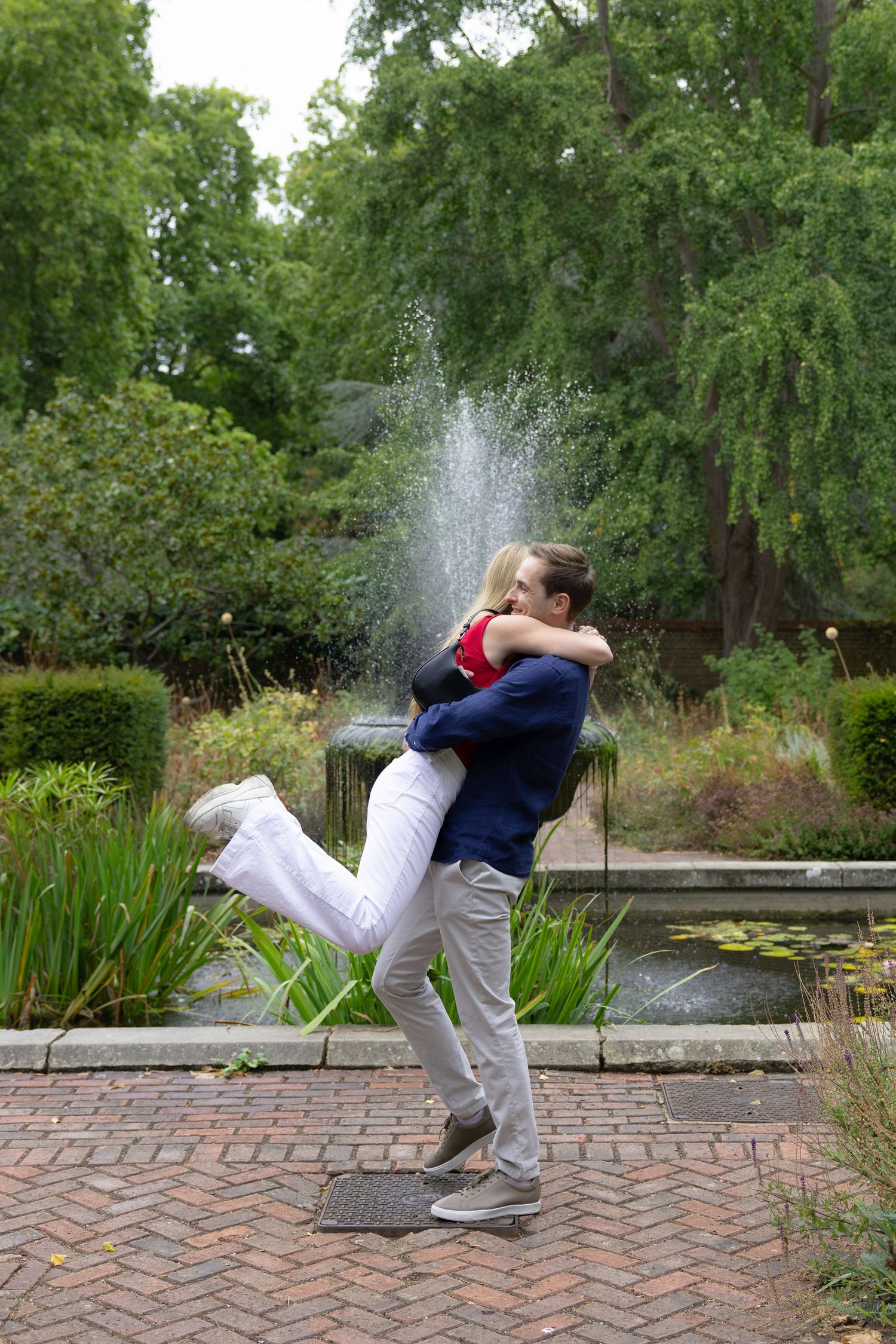 Couple sharing a romantic embrace by a garden fountain on a brick path surrounded by lush green trees.
