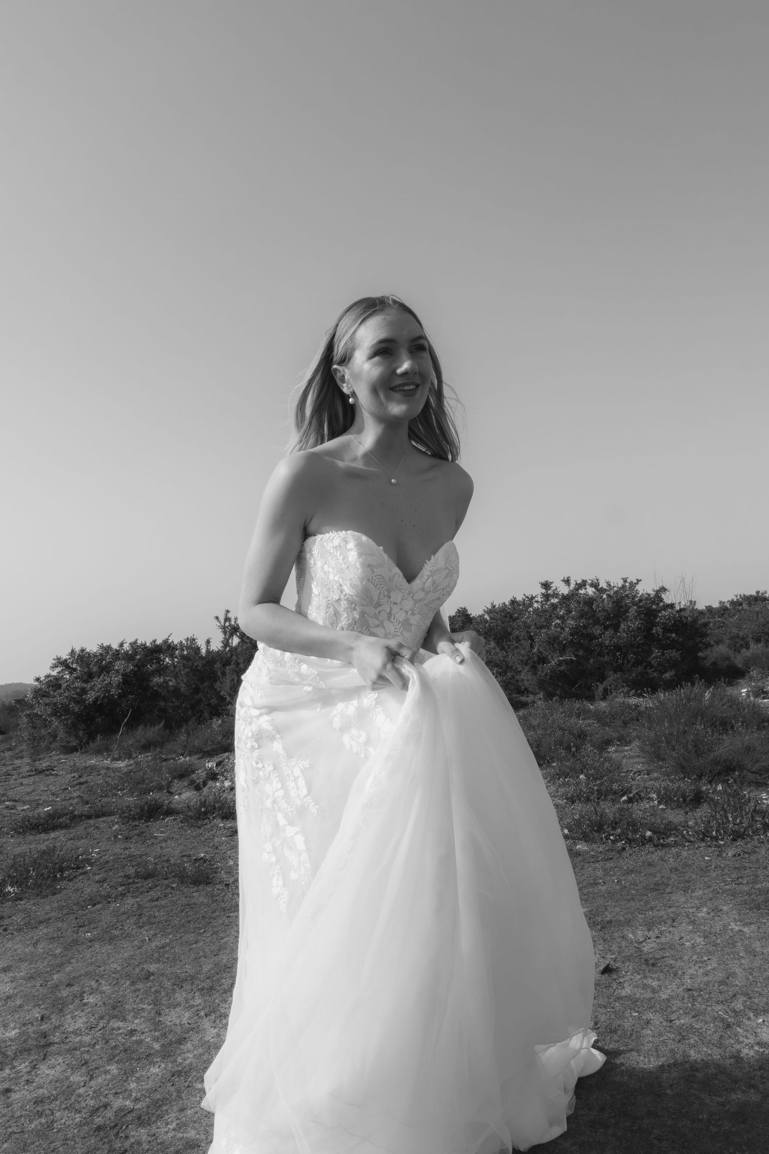 Smiling bride in strapless lace wedding dress posing outdoors in black and white photo.
