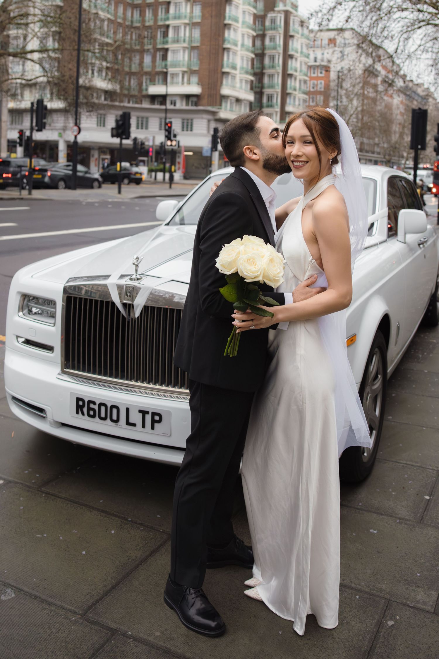 Bride and groom kissing beside a white Rolls-Royce on a London street, bride holding white roses bouquet.