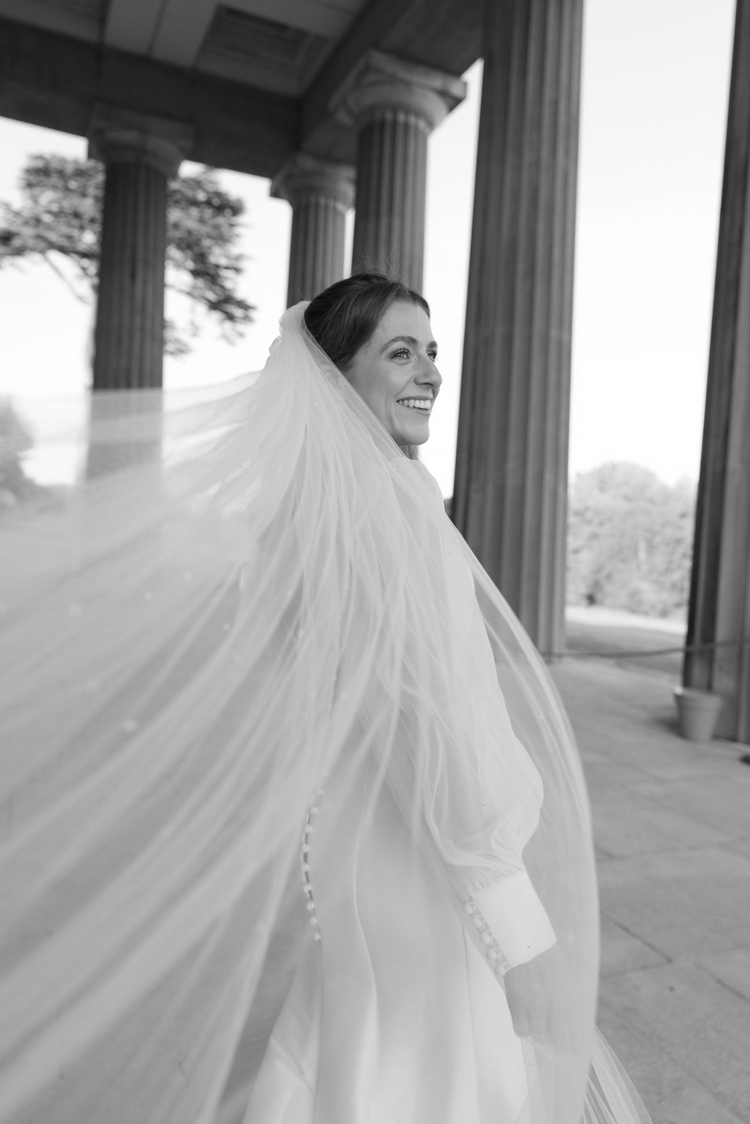 Dramatic black and white photo of flowing bridal veil and dress among classical pillars.