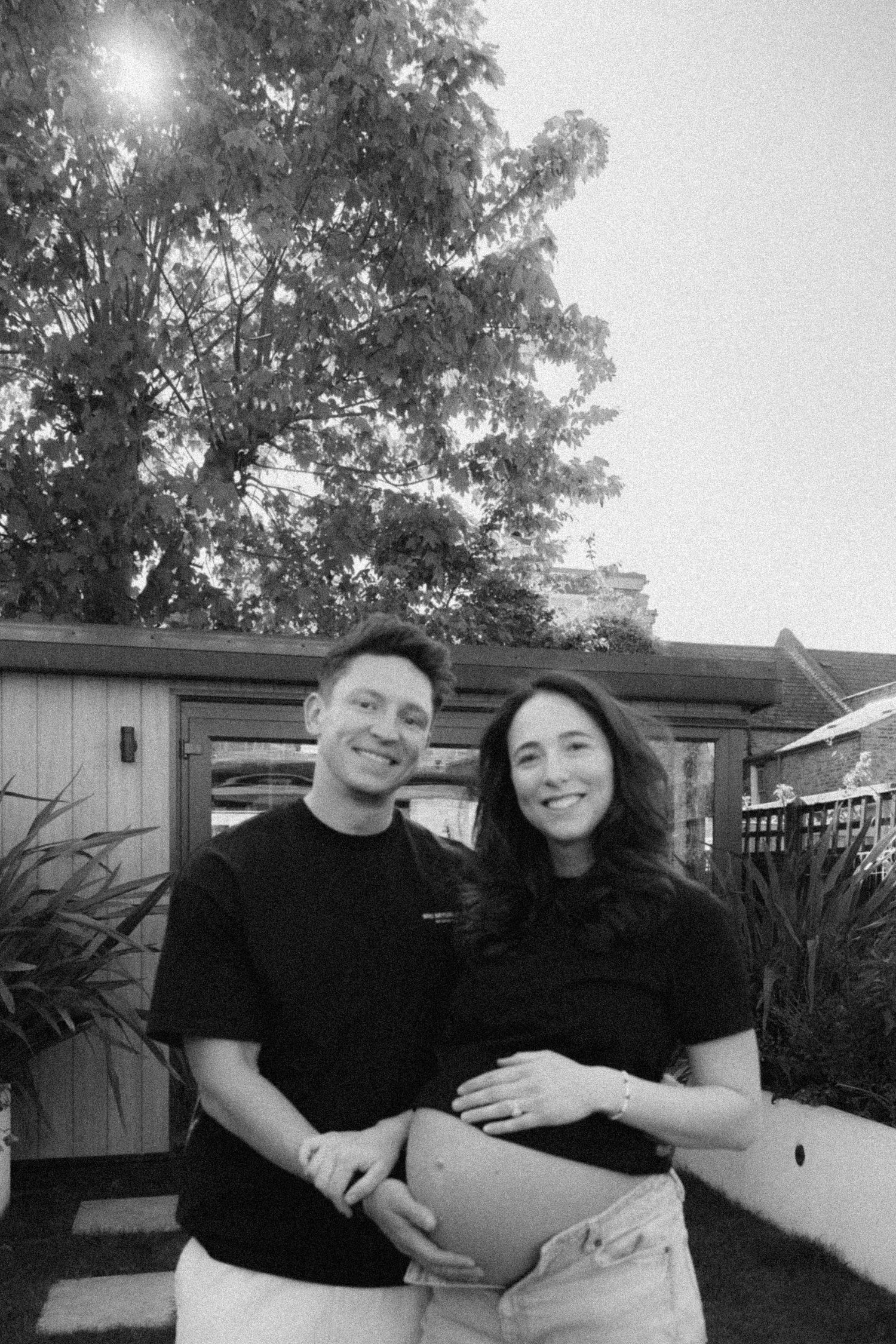 Black and white photo of smiling couple outdoors, woman showing baby bump, standing near garden.
