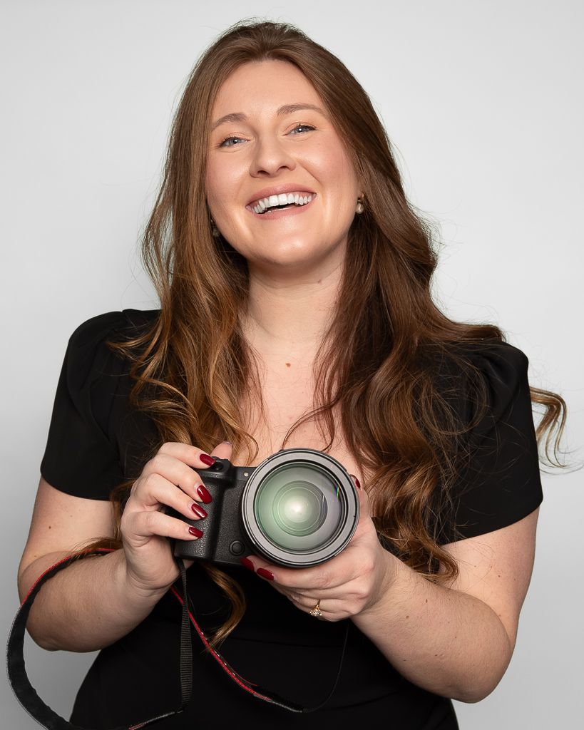 Professional photographer in black shirt smiling while holding a DSLR camera against white background.