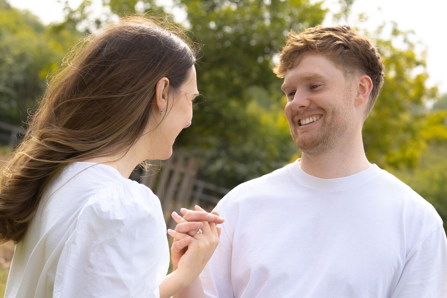 Happy couple holding hands outdoors, smiling at each other in a sunny green park setting.