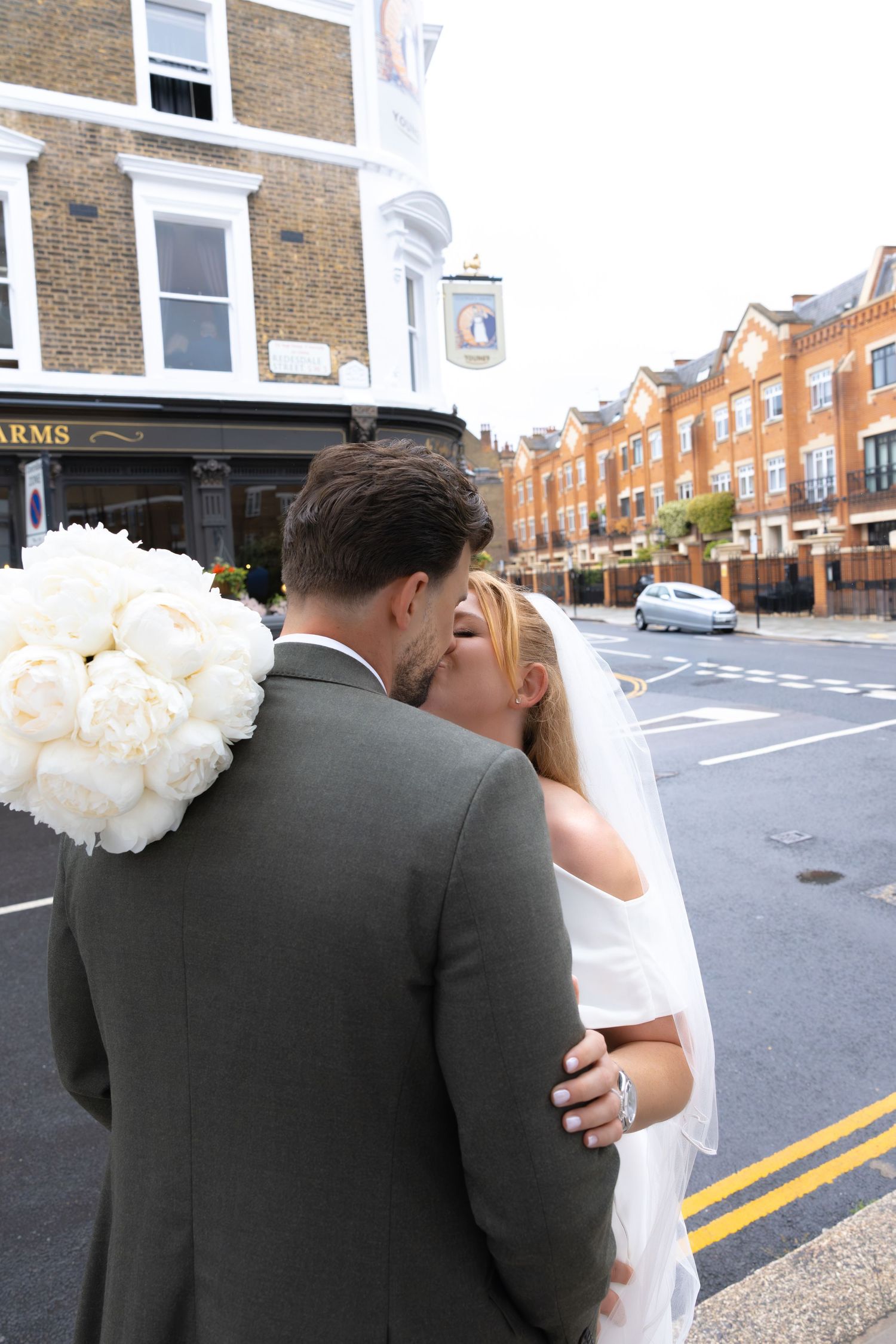 Bride and groom kissing on a London street, bride holding white peony bouquet wearing veil, groom in grey suit.