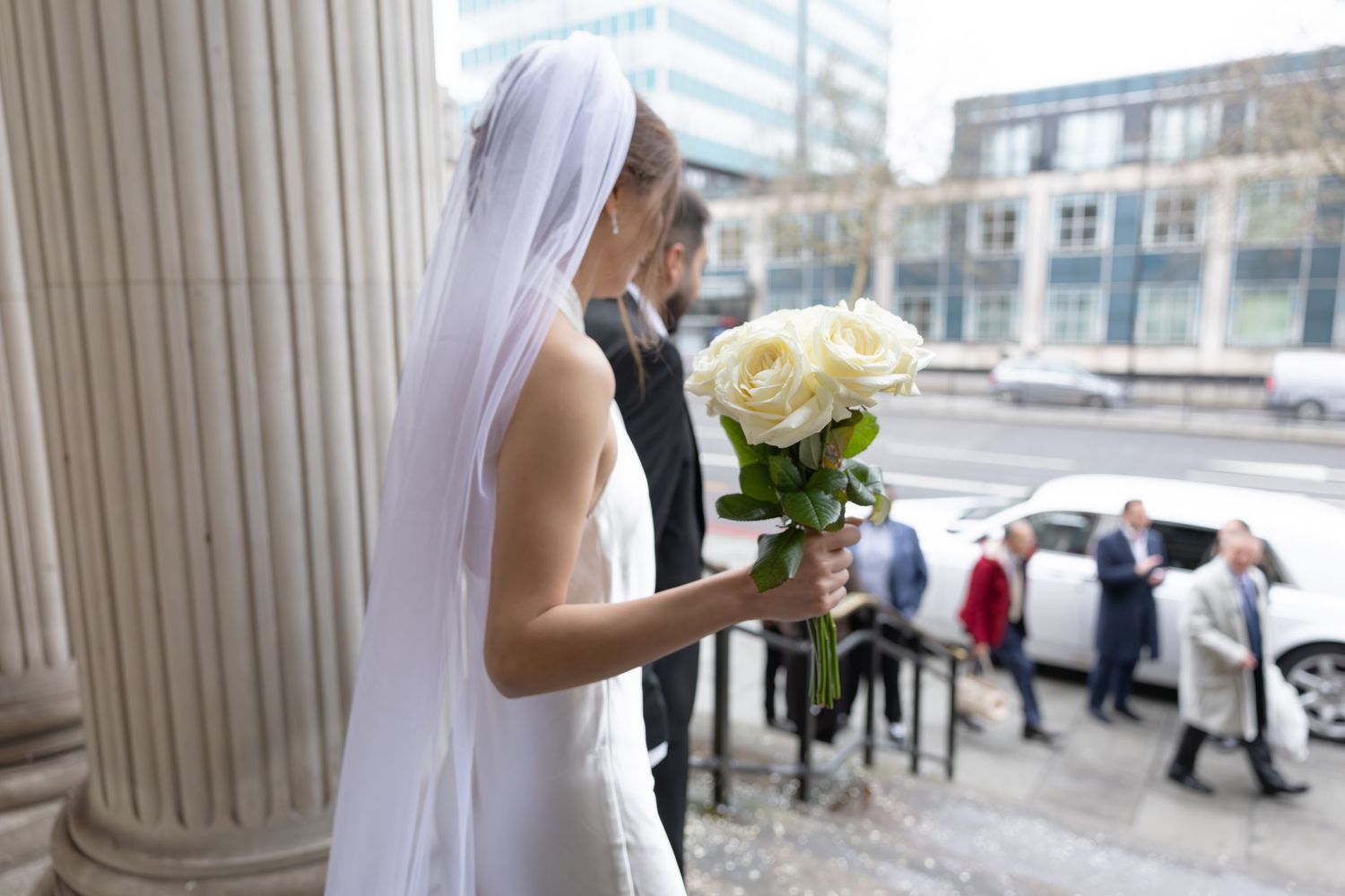Bride holding white rose bouquet stands by stone column outside wedding venue on city street.