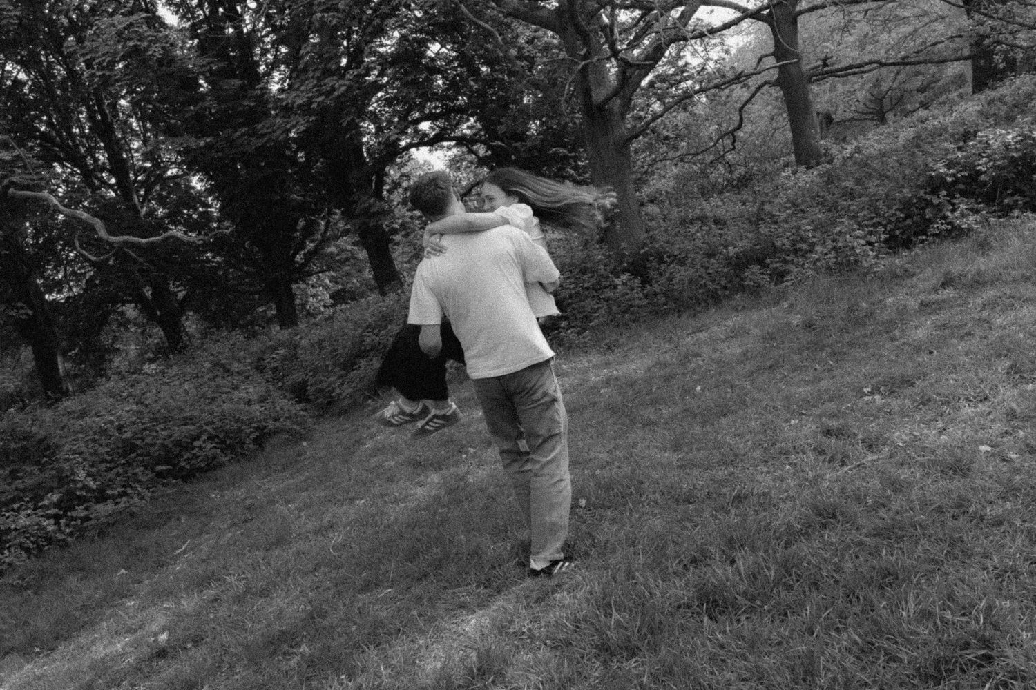 Black and white photo of a person carrying a child on their back in a lush green park setting.