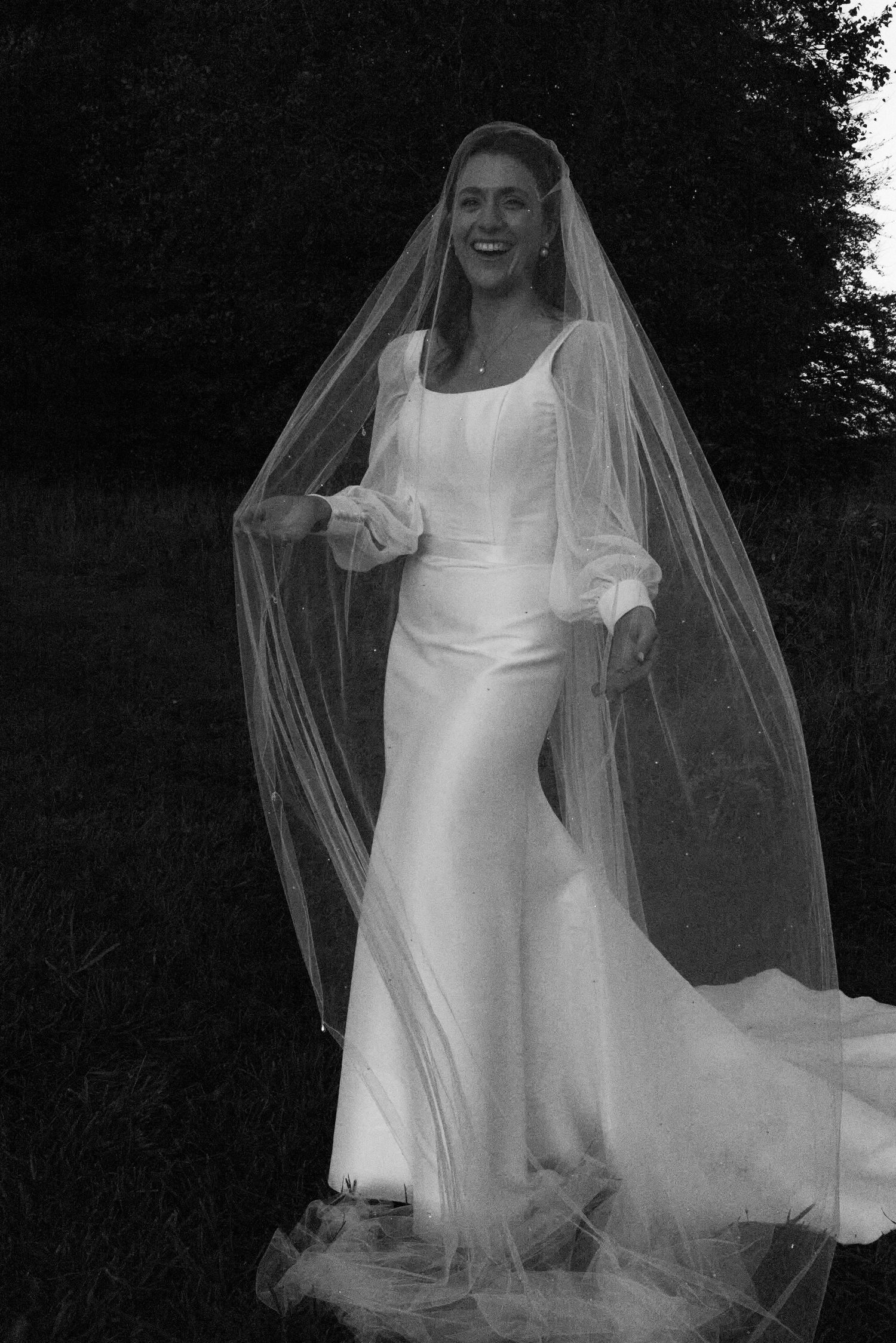 Bride in an elegant white dress with flowing veil against a dark background.