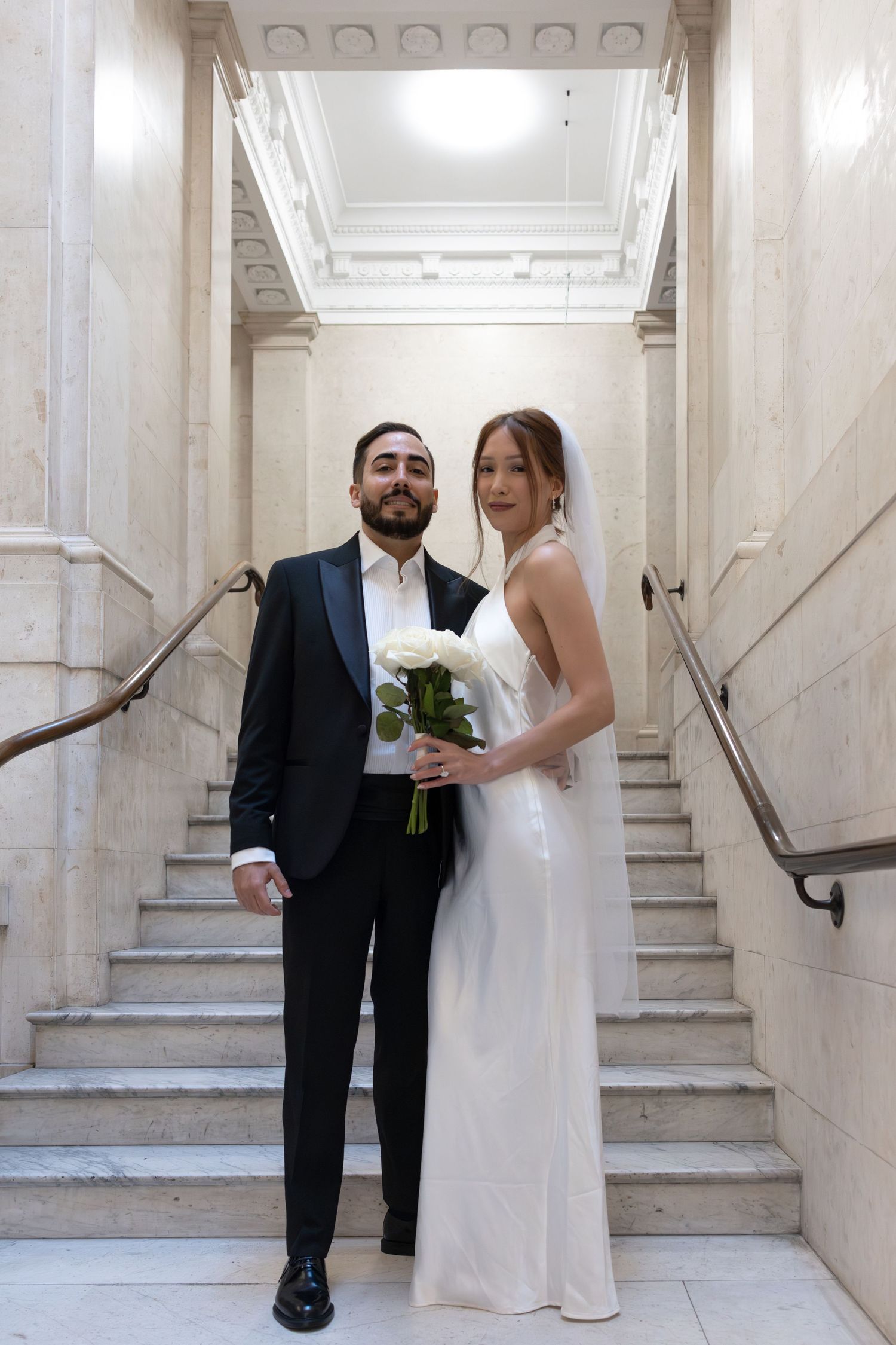 Newlywed couple poses on marble staircase, bride in white dress with veil holding white roses, groom in black tuxedo.
