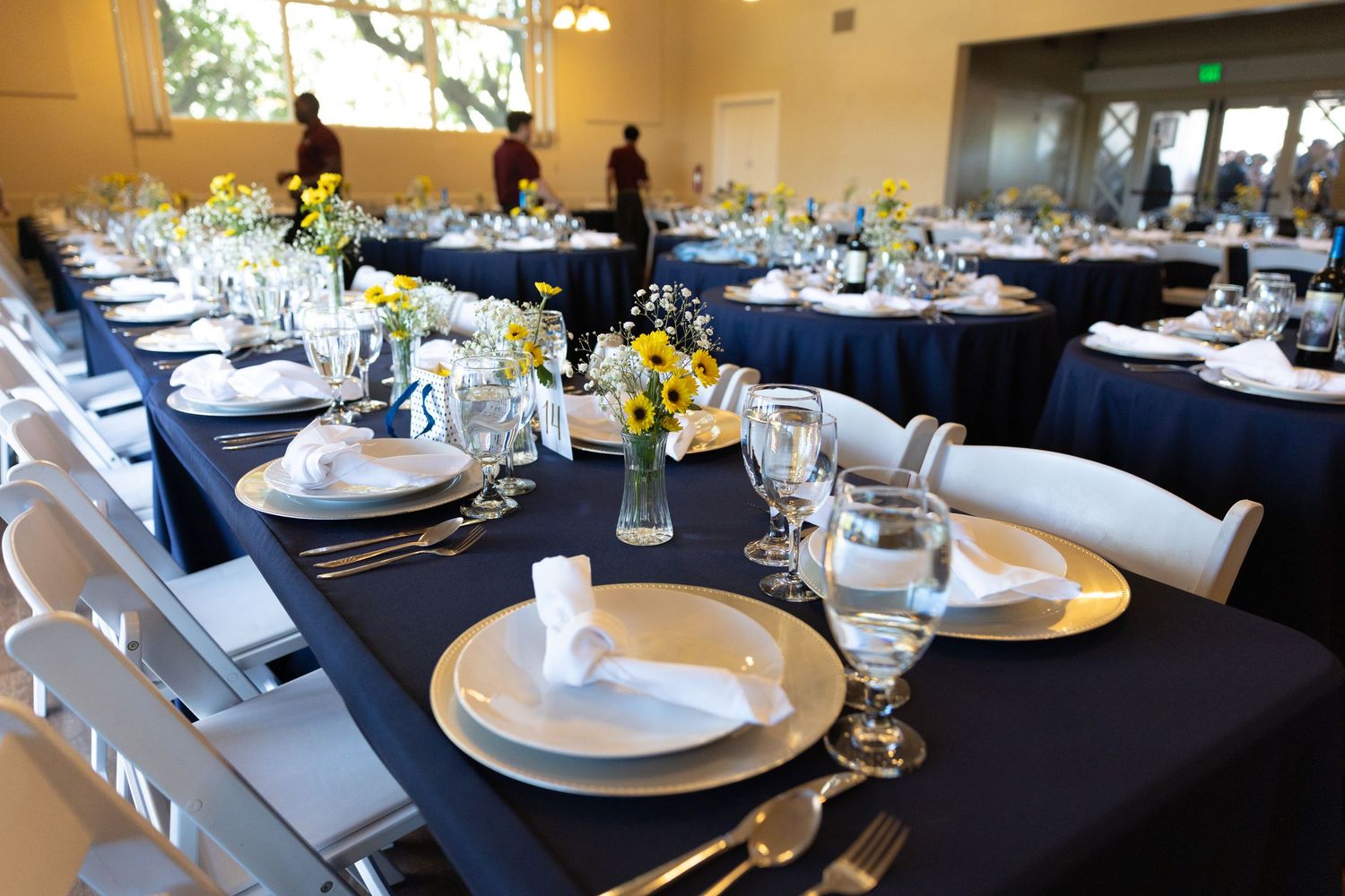 Elegant wedding reception tables with navy blue linens, white place settings, and yellow floral centerpieces in a bright venue.