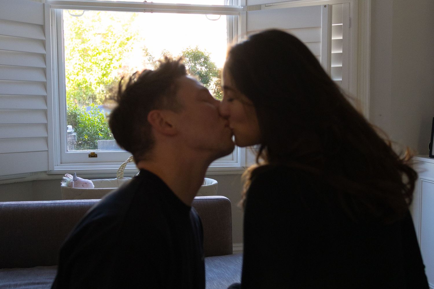 Couple sharing a romantic kiss in silhouette near a bright window indoors.