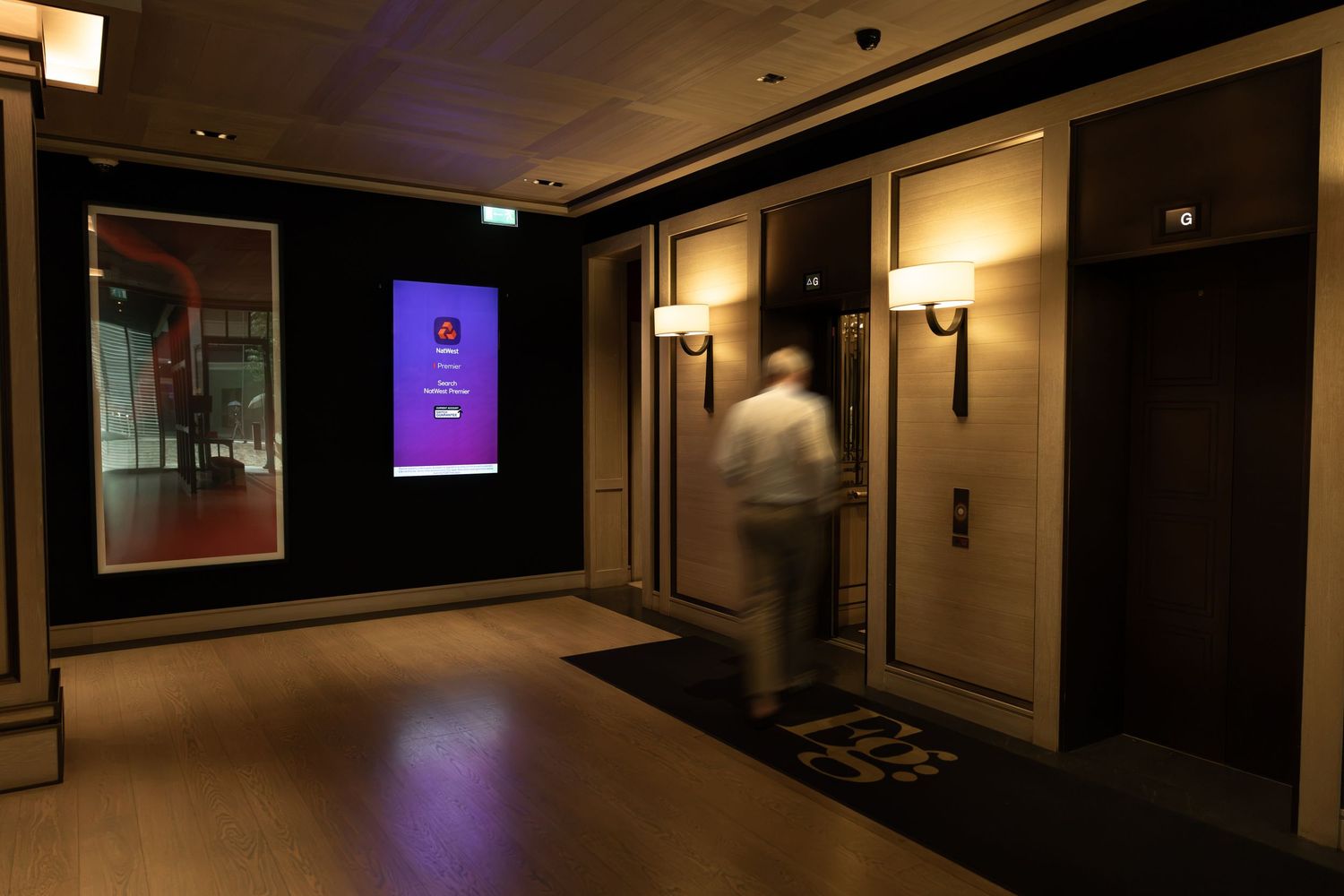 A blurred figure walks past elevator doors in a modern hotel lobby with warm lighting and a digital display screen.