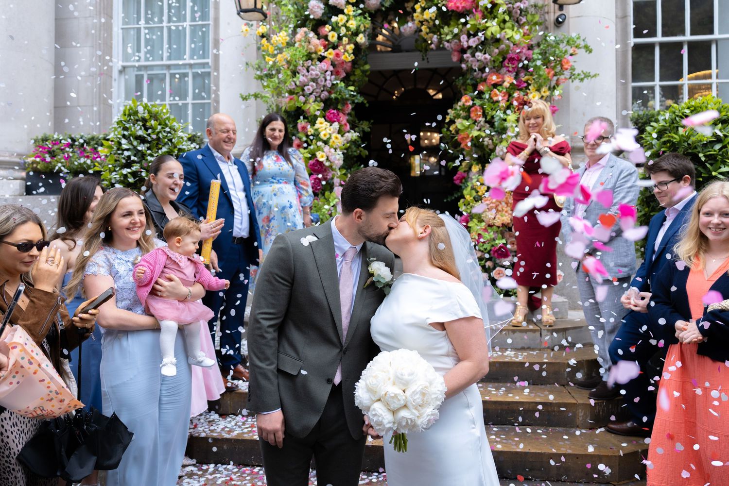 Newlyweds kissing on steps as guests throw colorful confetti outside a flower-adorned venue.