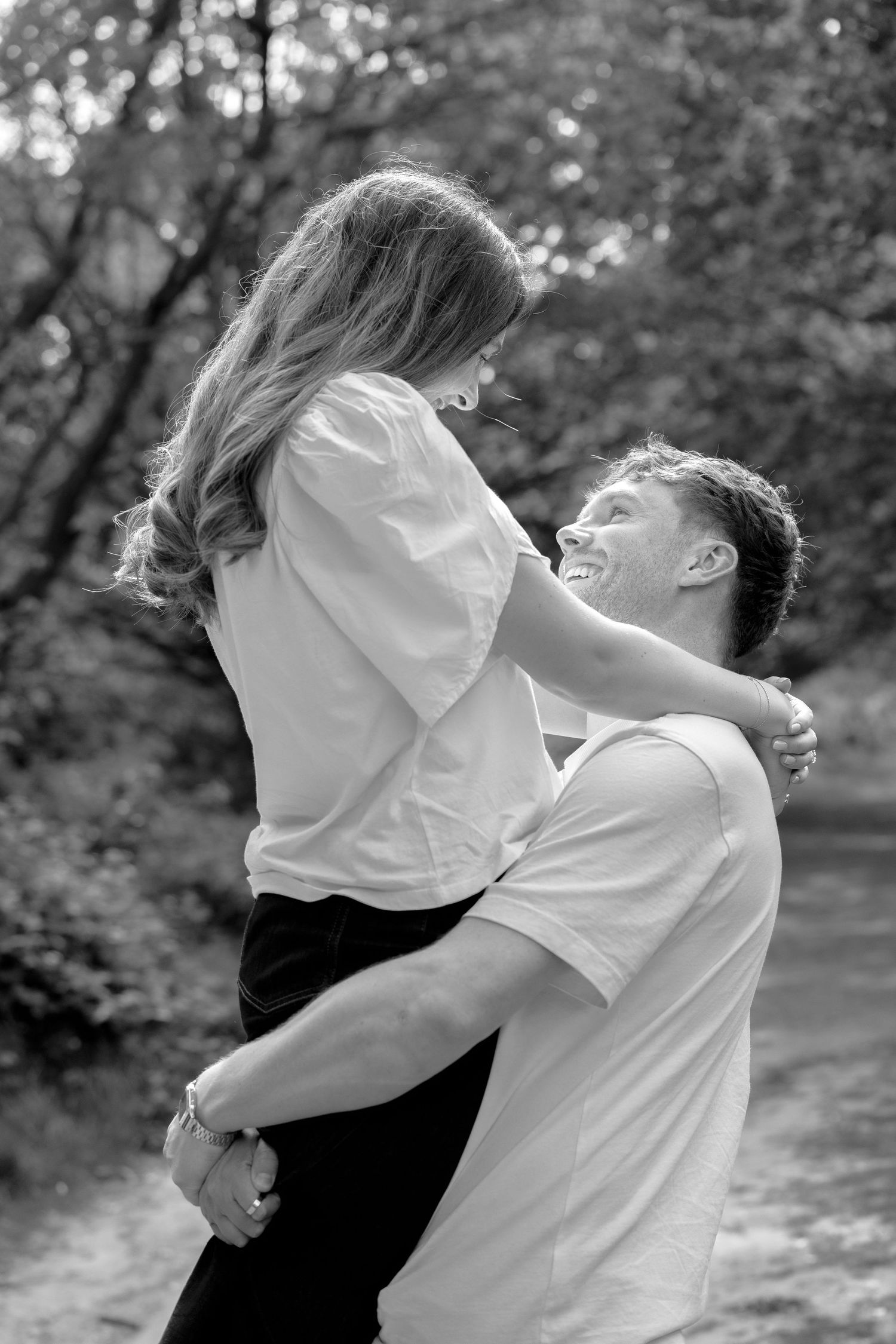 Black and white photo of a man lifting and smiling at a woman outdoors, capturing a joyful romantic moment.