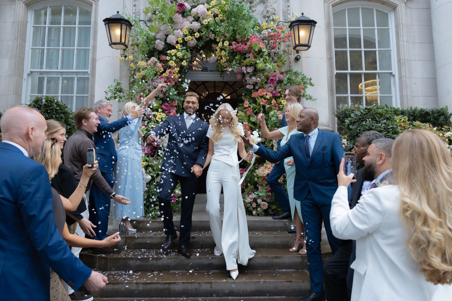 Bride and groom exit venue on flower-adorned steps as guests throw confetti during joyful outdoor wedding celebration.