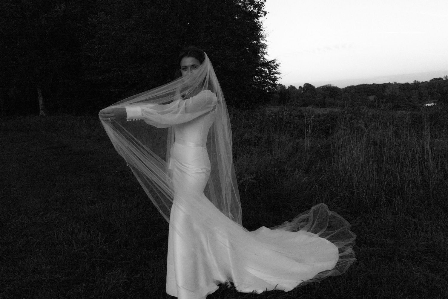 Silhouetted bride in flowing veil poses against a darkening landscape.