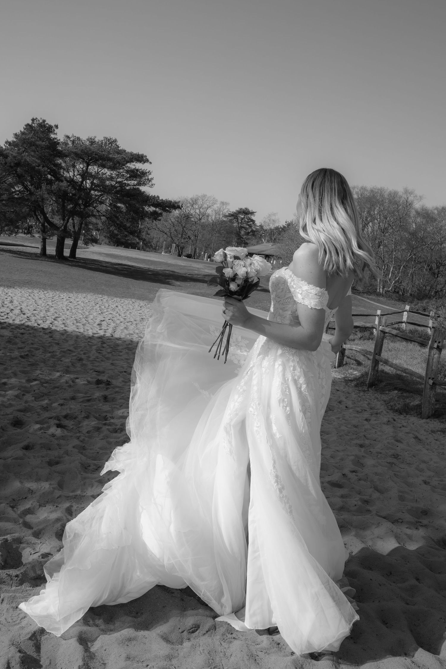 Black and white photo of a bride in a flowing off-shoulder gown holding a bouquet on a sandy beach.