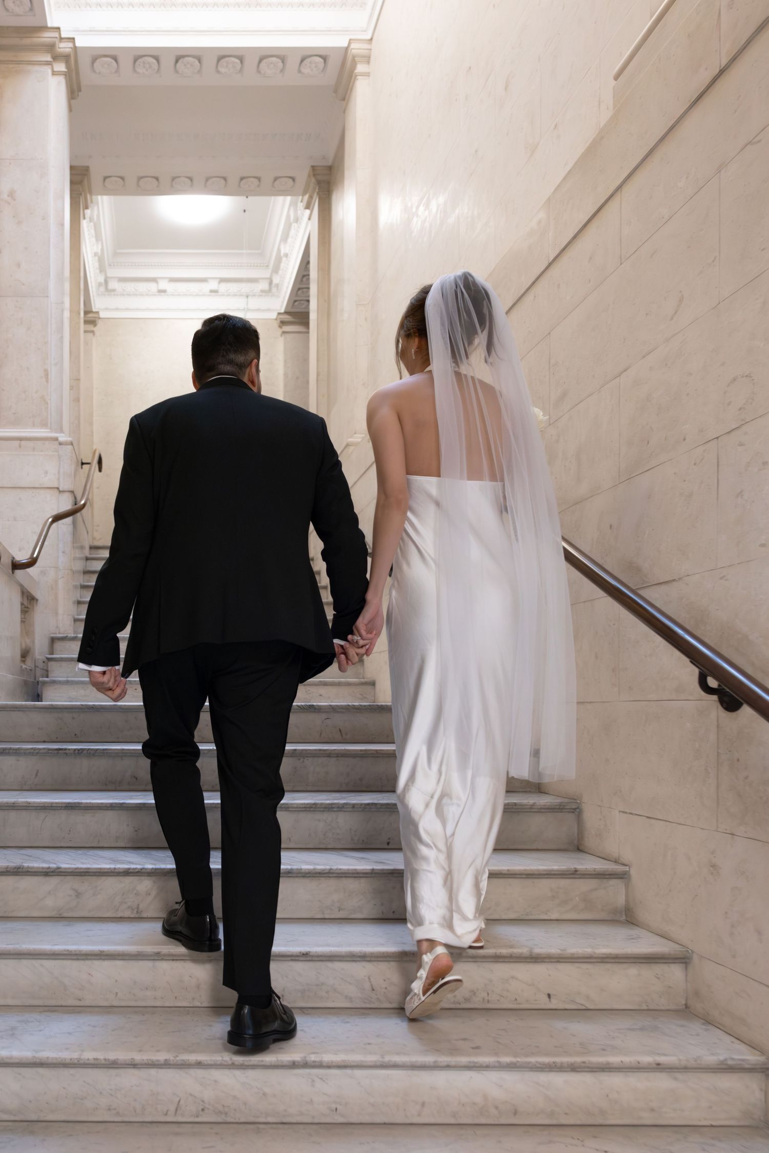 Bride and groom walking up marble stairs hand in hand, bride wearing white gown and veil, groom in black suit.