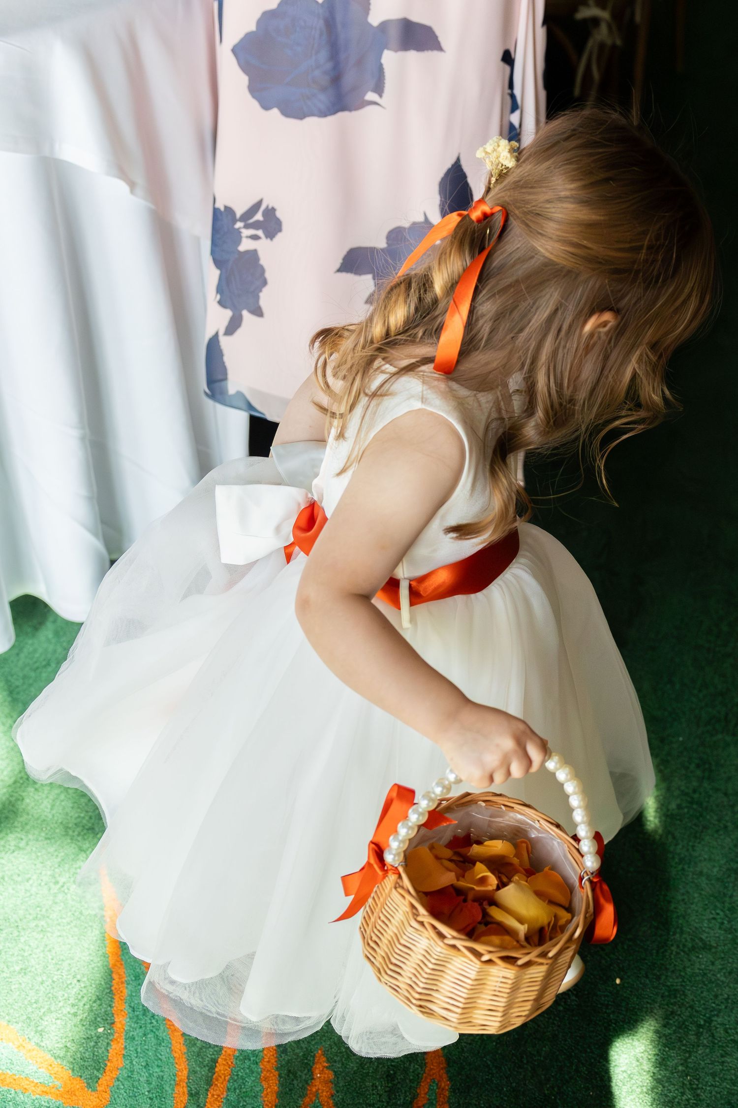 A child in a white dress with orange ribbons holds a wicker basket while standing on green carpet.