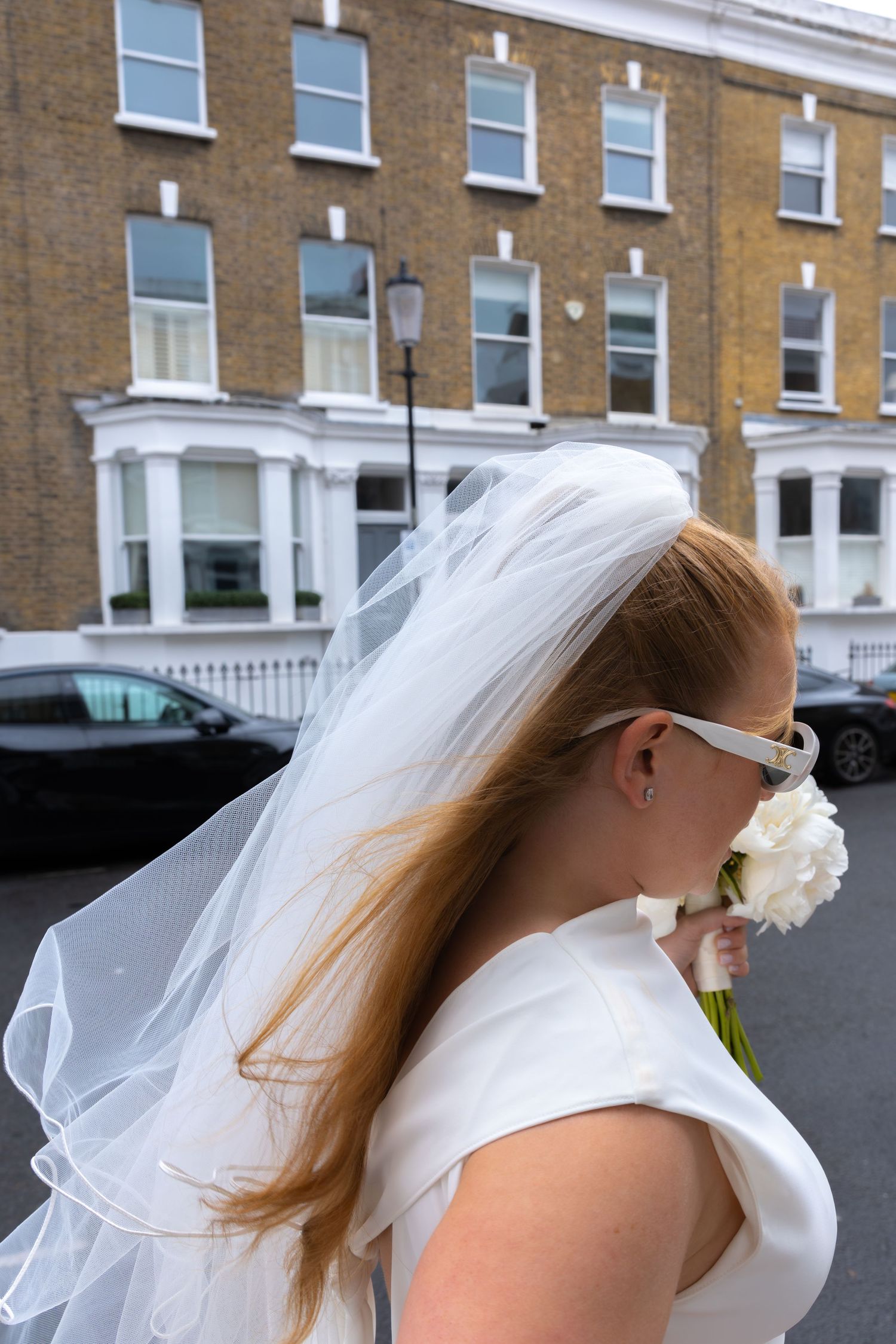 Red-haired bride in white dress and veil smells white bouquet outside a London brick building.