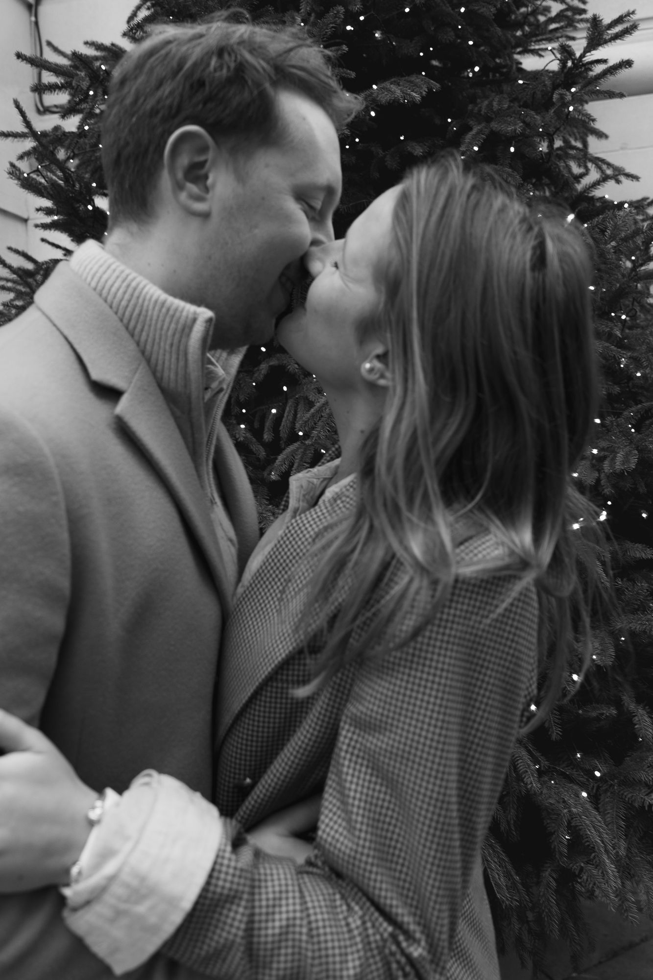 A couple shares a romantic moment by a decorated Christmas tree with twinkling lights.