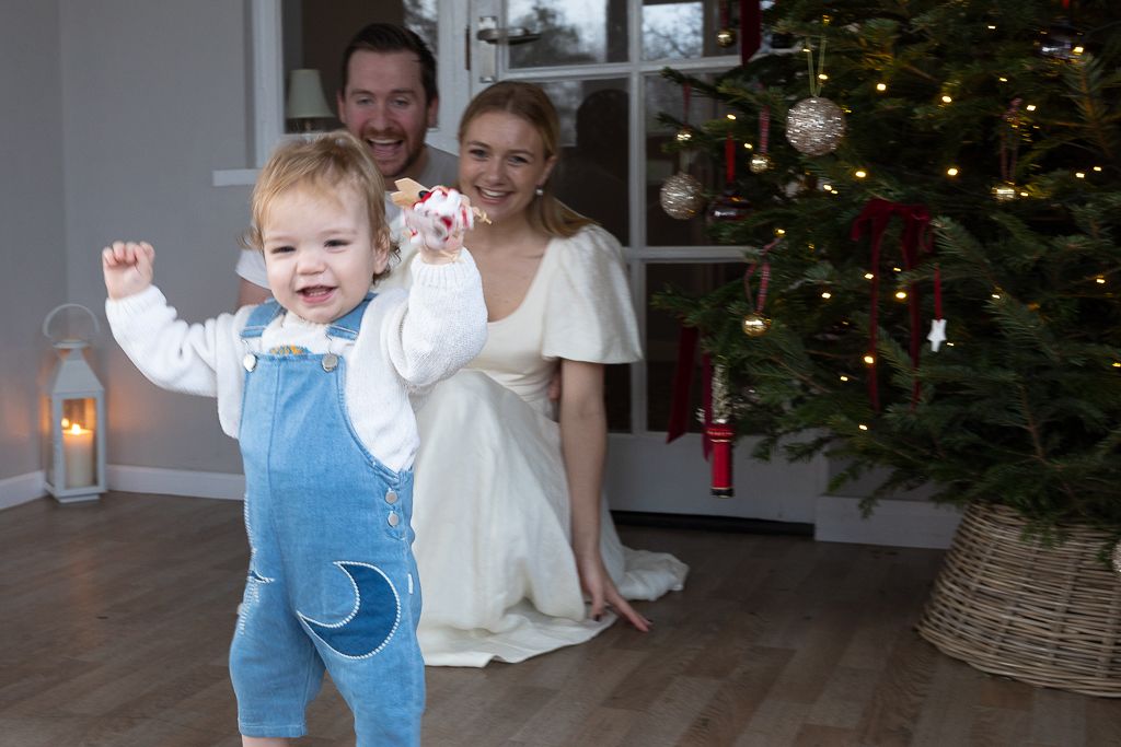 A festive indoor scene with a decorated Christmas tree mum, dad, and toddler celebrate the toddlers first steps.