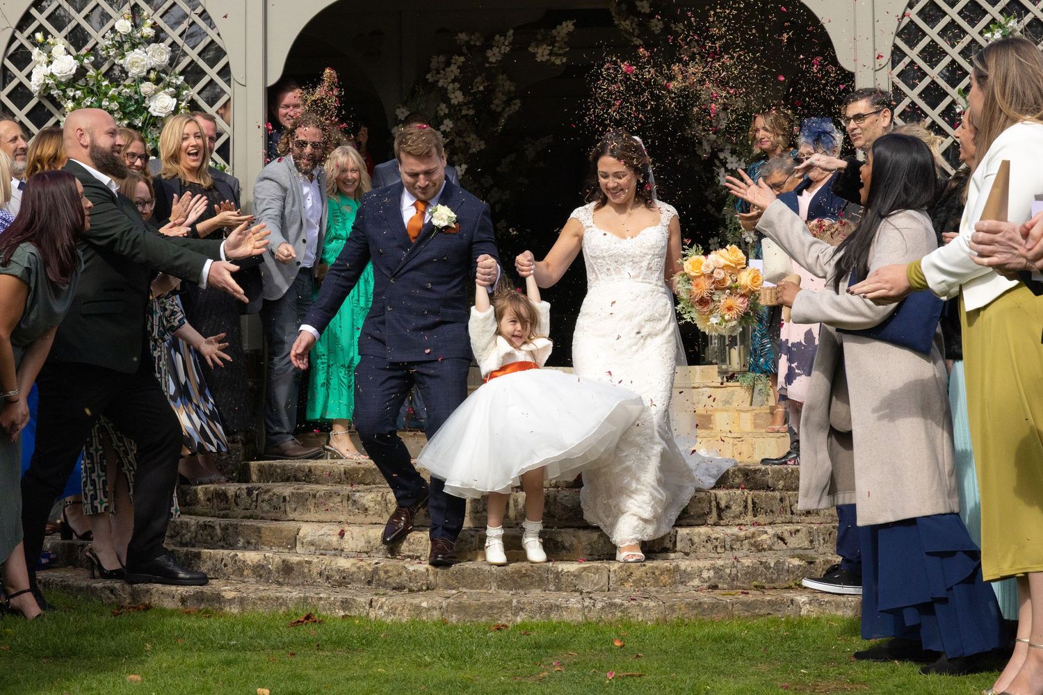 A group of wedding guests gather outdoors near a stone wall to take photos at a garden reception.