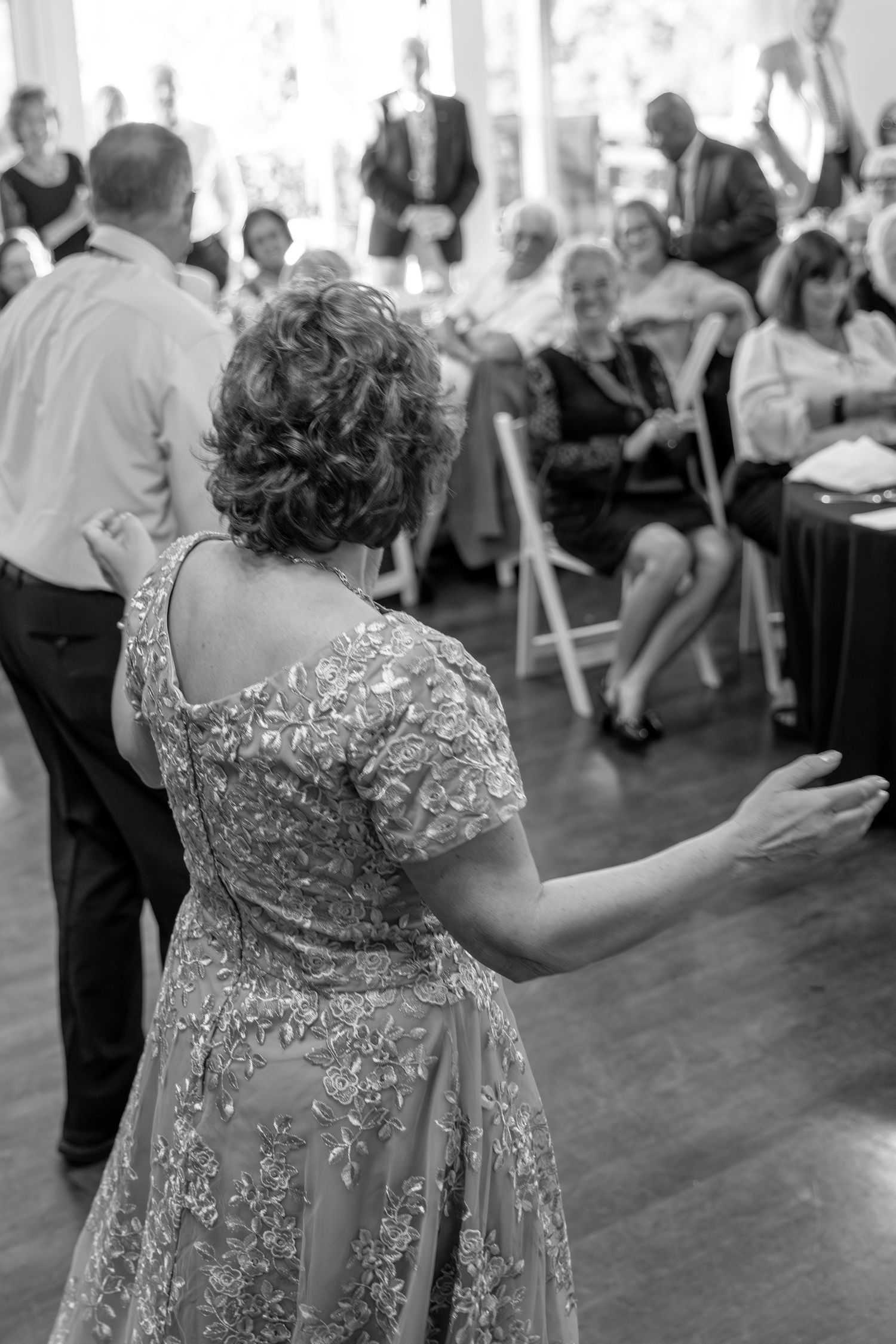 Black and white photo of a woman in a floral dress dancing at a wedding reception with guests seated in background.