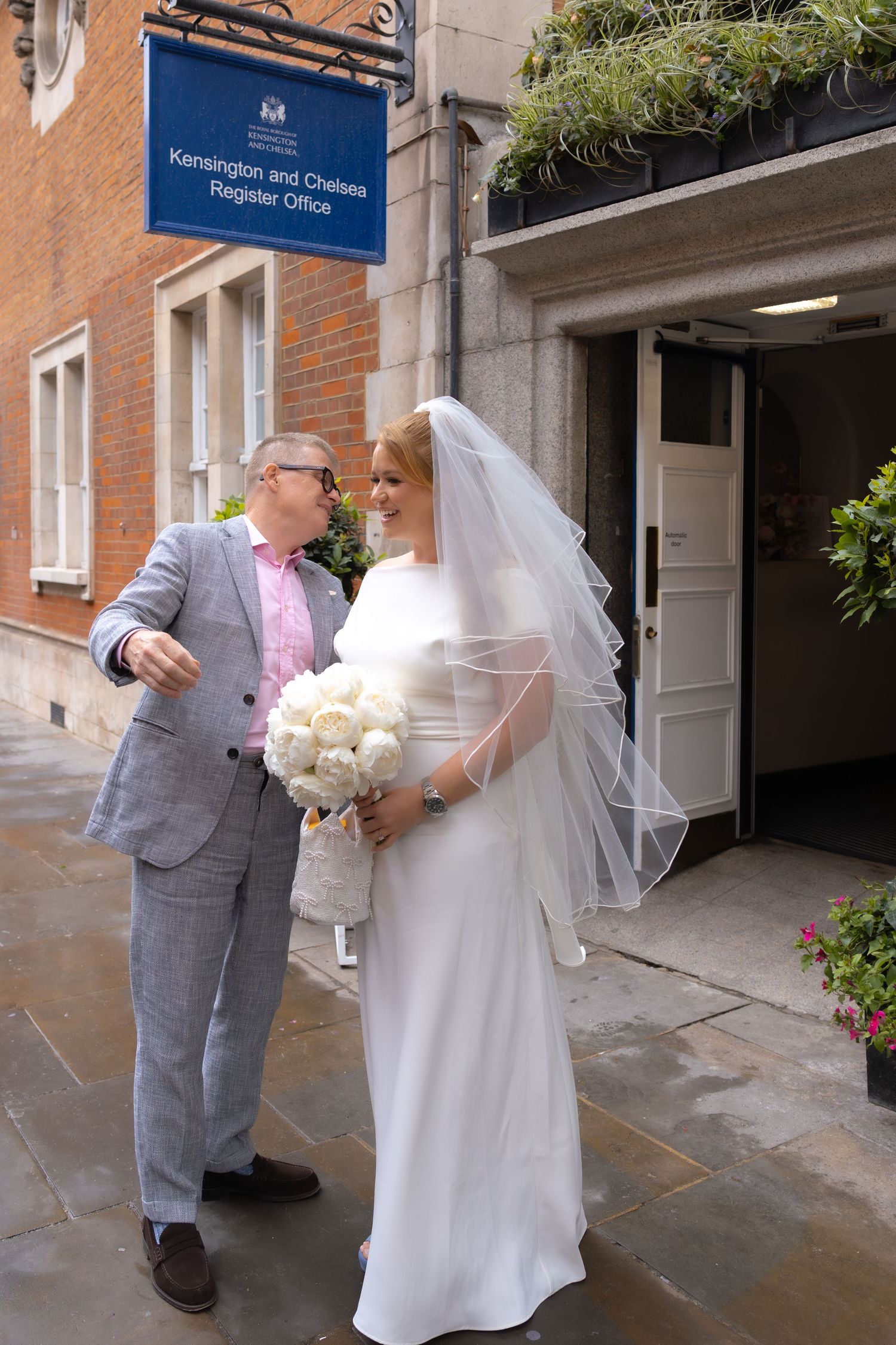 Bride and groom outside Kensington and Chelsea Register Office, bride holding white bouquet.