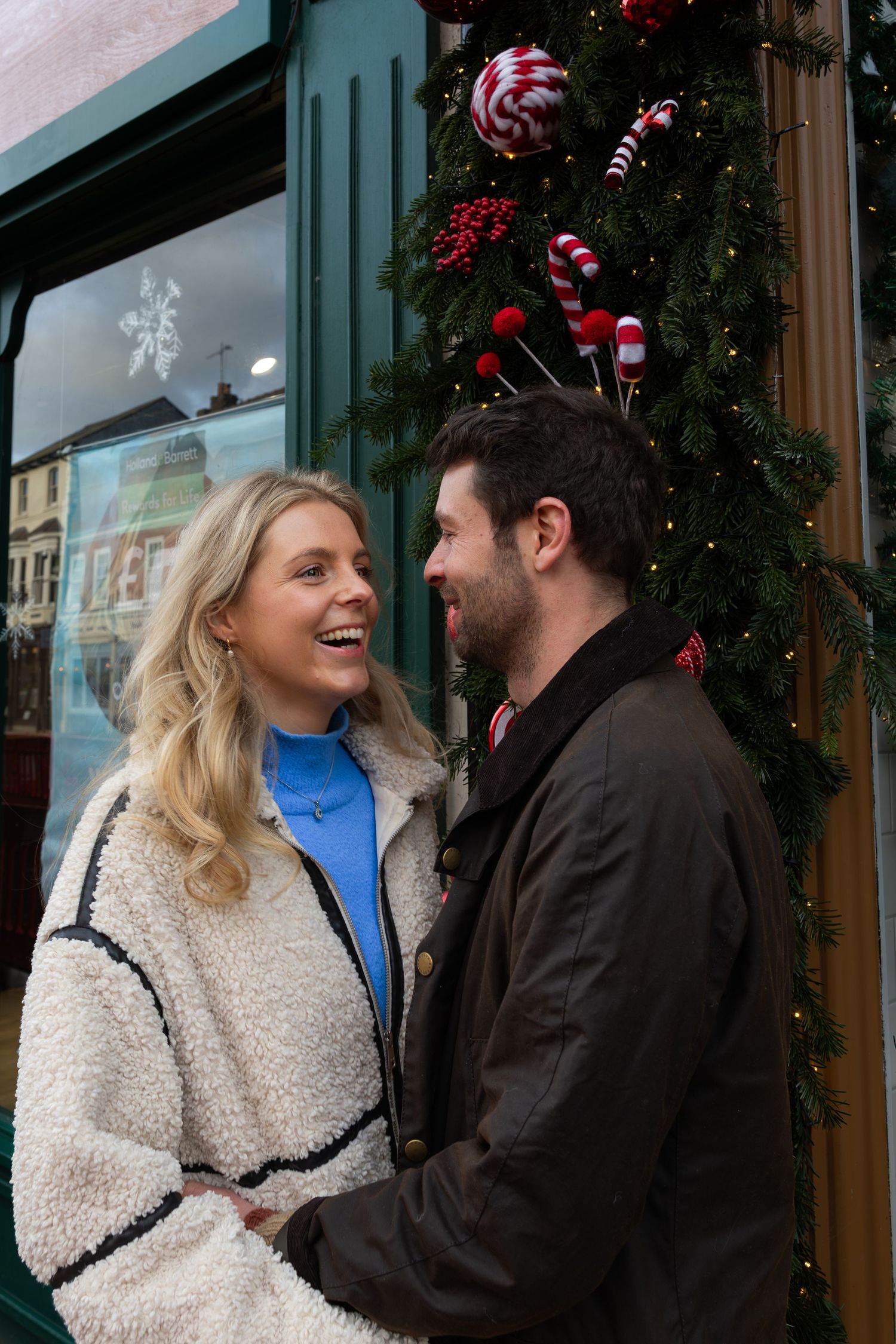 A couple laughs together in front of a storefront decorated with festive holiday garland and lights.