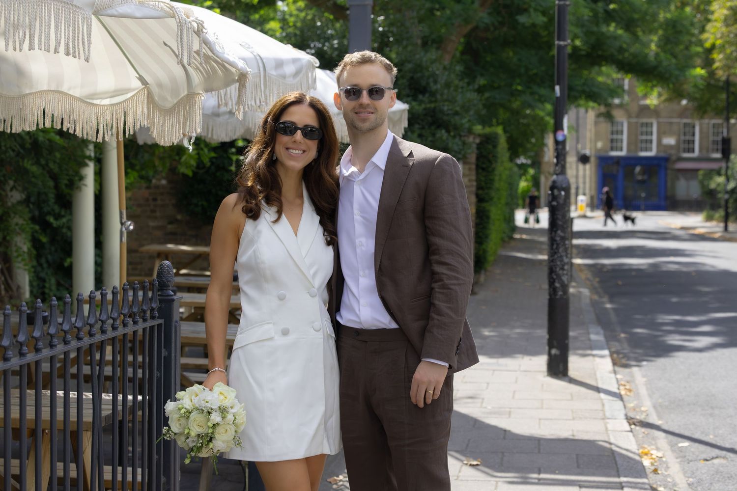 Couple in wedding attire posing on a sunny London street, woman in white dress holding bouquet, man in brown suit.