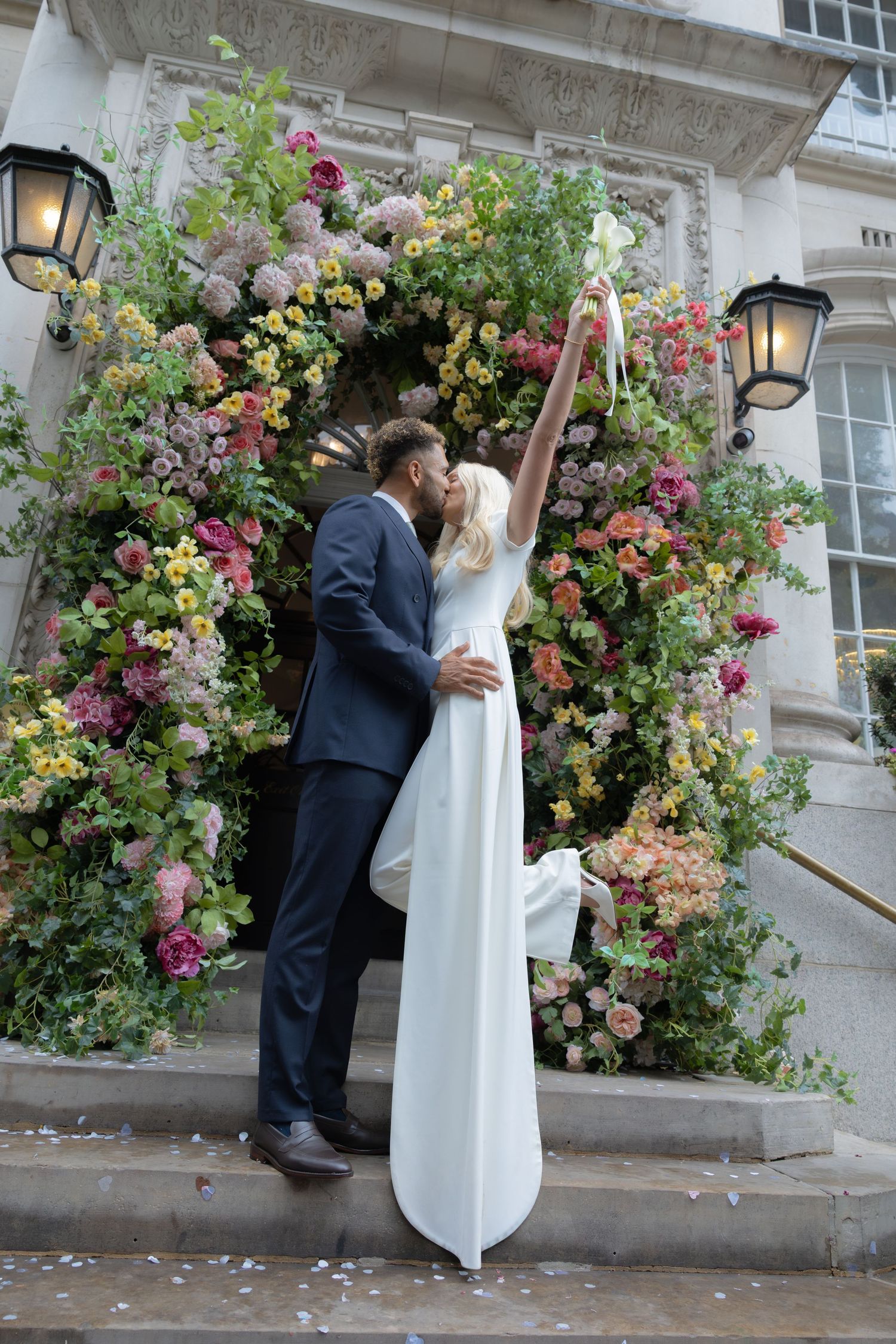 Bride and groom kissing on steps before a colorful floral arch at an elegant wedding venue.