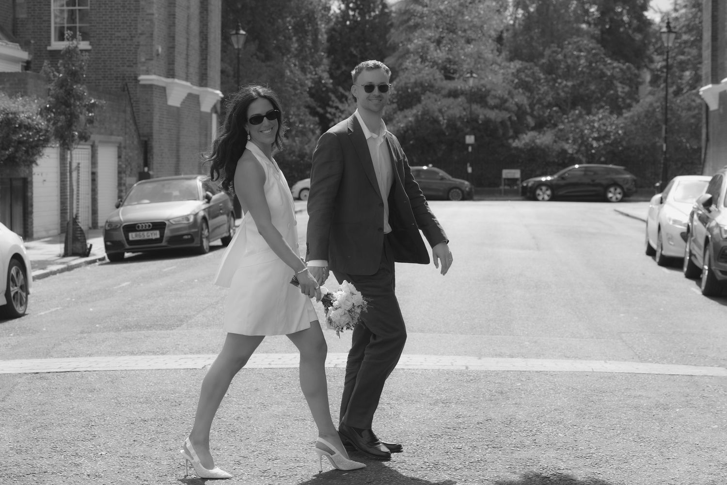 Black and white photo of a bride and groom holding hands crossing a street, bride in short dress holding bouquet.