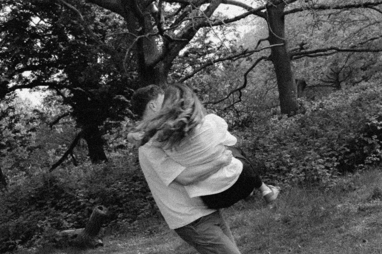 Black and white photo of two people embracing and spinning outdoors among trees.