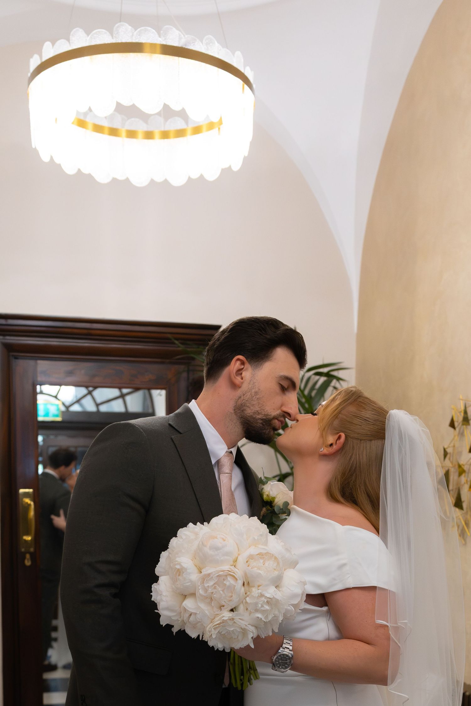 Bride and groom sharing a romantic kiss indoors, bride holding white peony bouquet, elegant chandelier overhead.