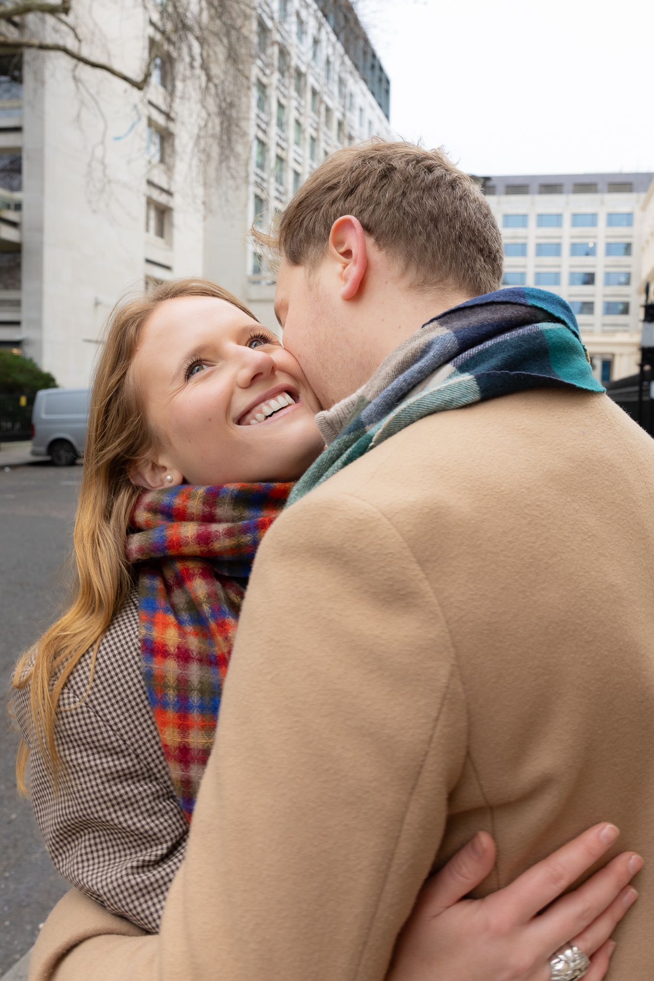 A couple in casual winter wear shares an affectionate moment outside historic buildings.
