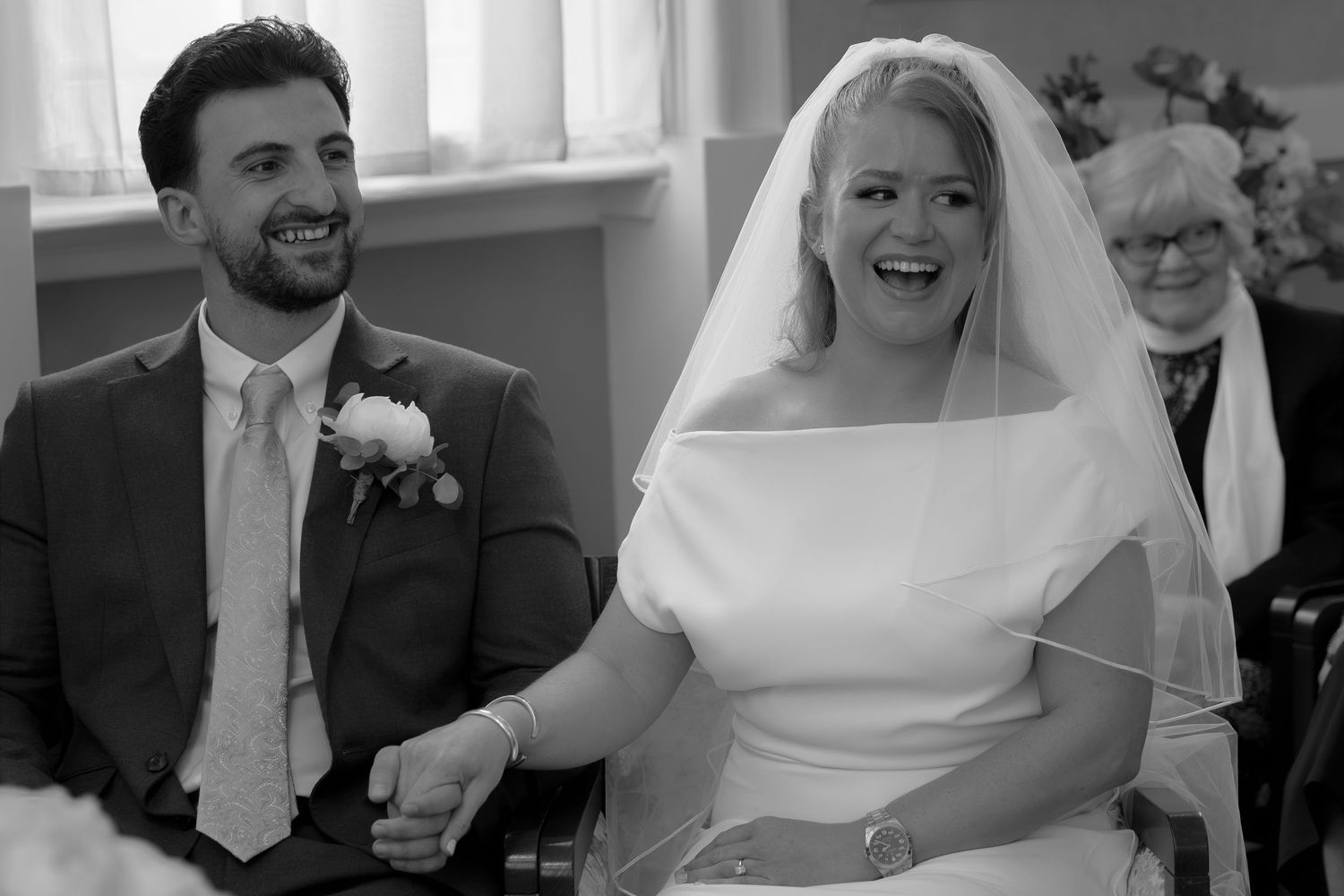 Black and white photo of a smiling bride in veil and groom in suit laughing together during wedding ceremony.