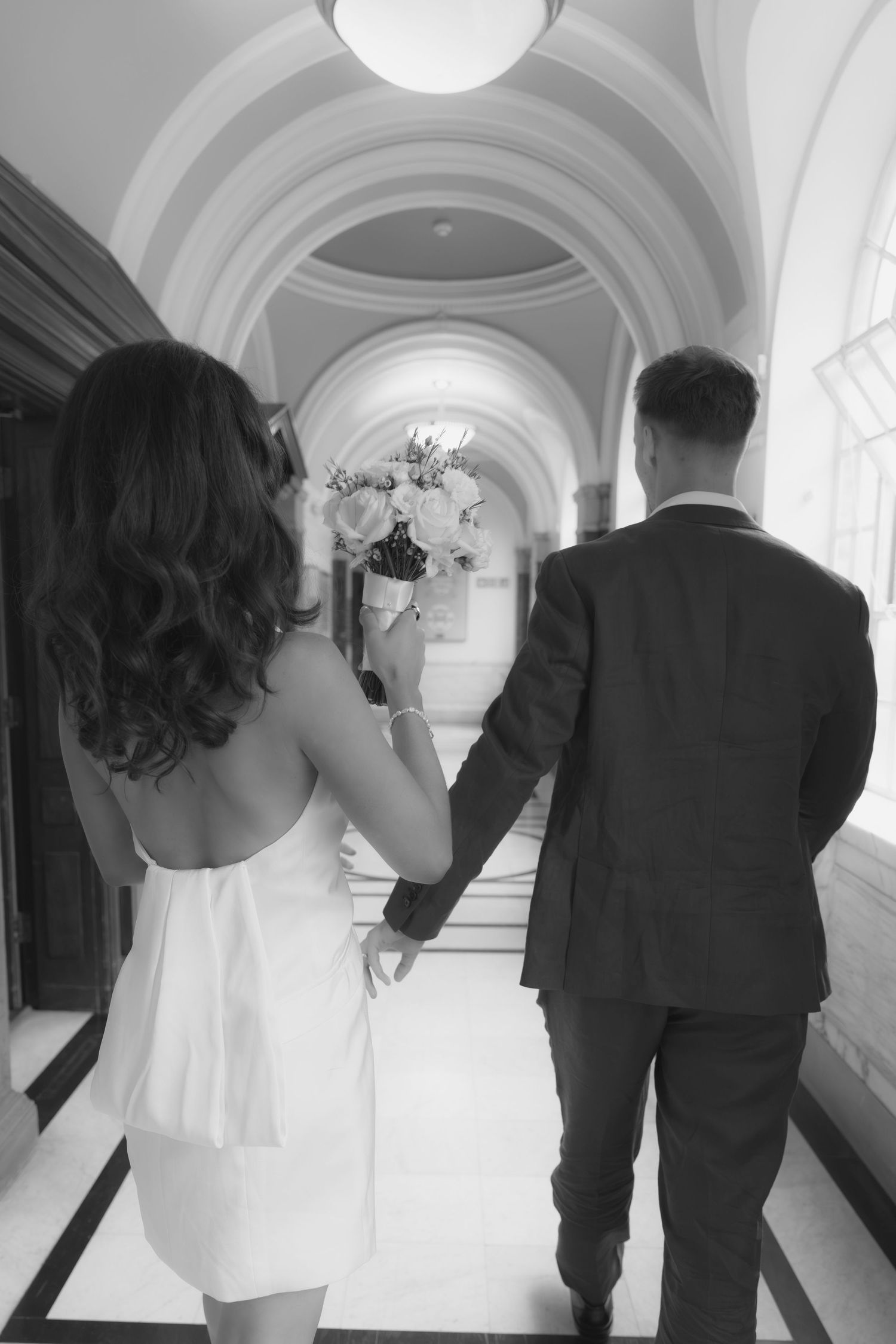 Black and white photo of a bride and groom holding hands walking down an elegant arched hallway, bride carrying a bouquet.