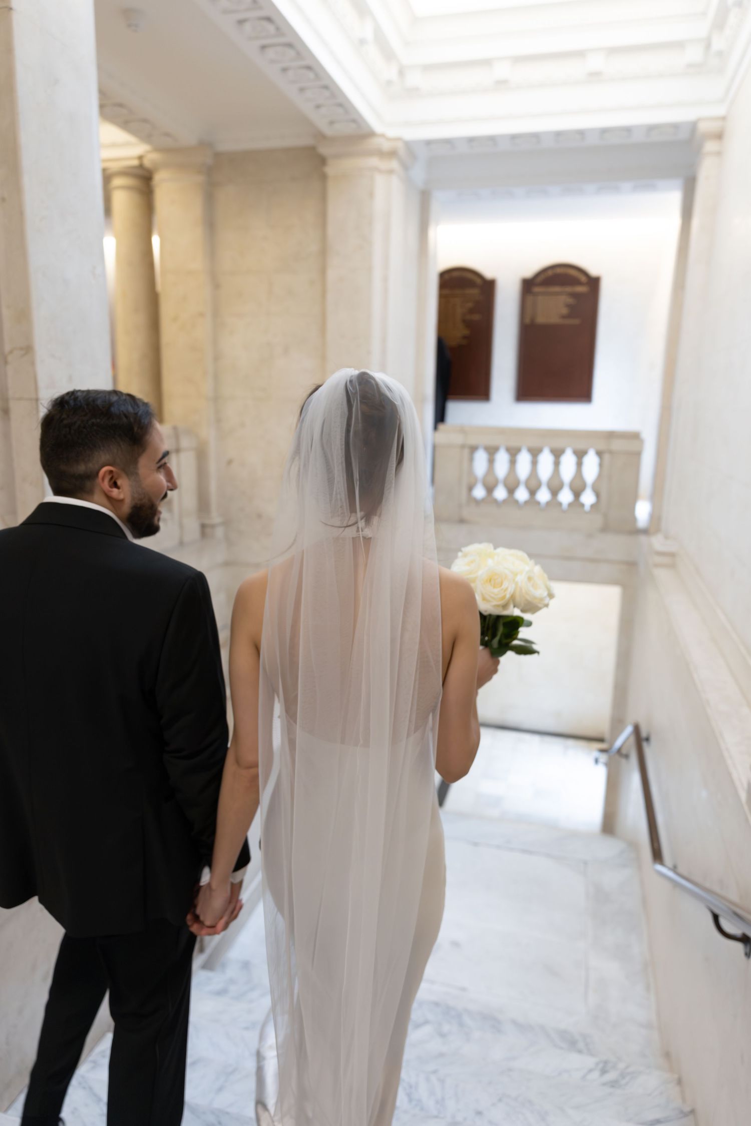 Bride holding white rose bouquet and groom descending marble staircase in elegant wedding venue.