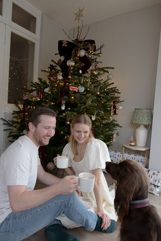 People sharing drinks and laughter by a decorated Christmas tree in a bright living room.
